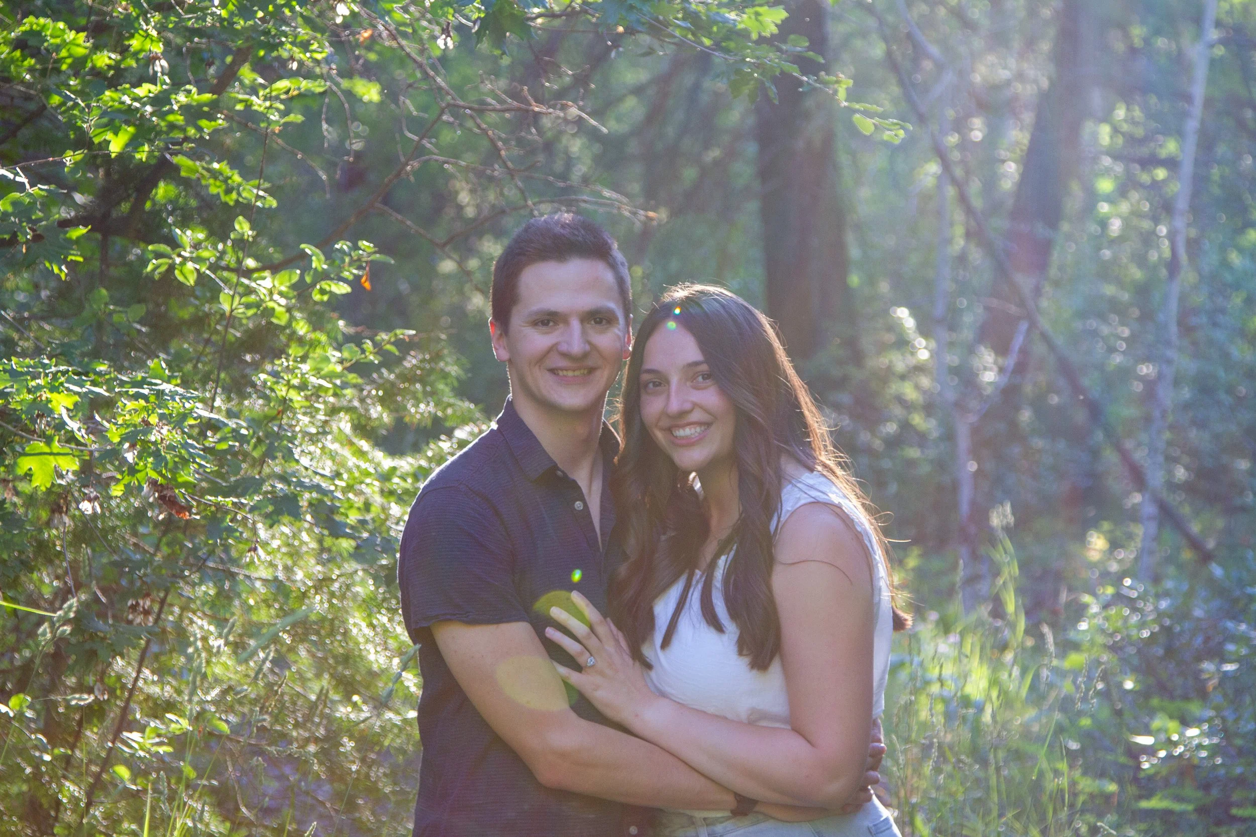 A smiling young man and woman standing closely together outdoors in a forest with sunlight filtering through trees, embracing each other with arms wrapped around each other.