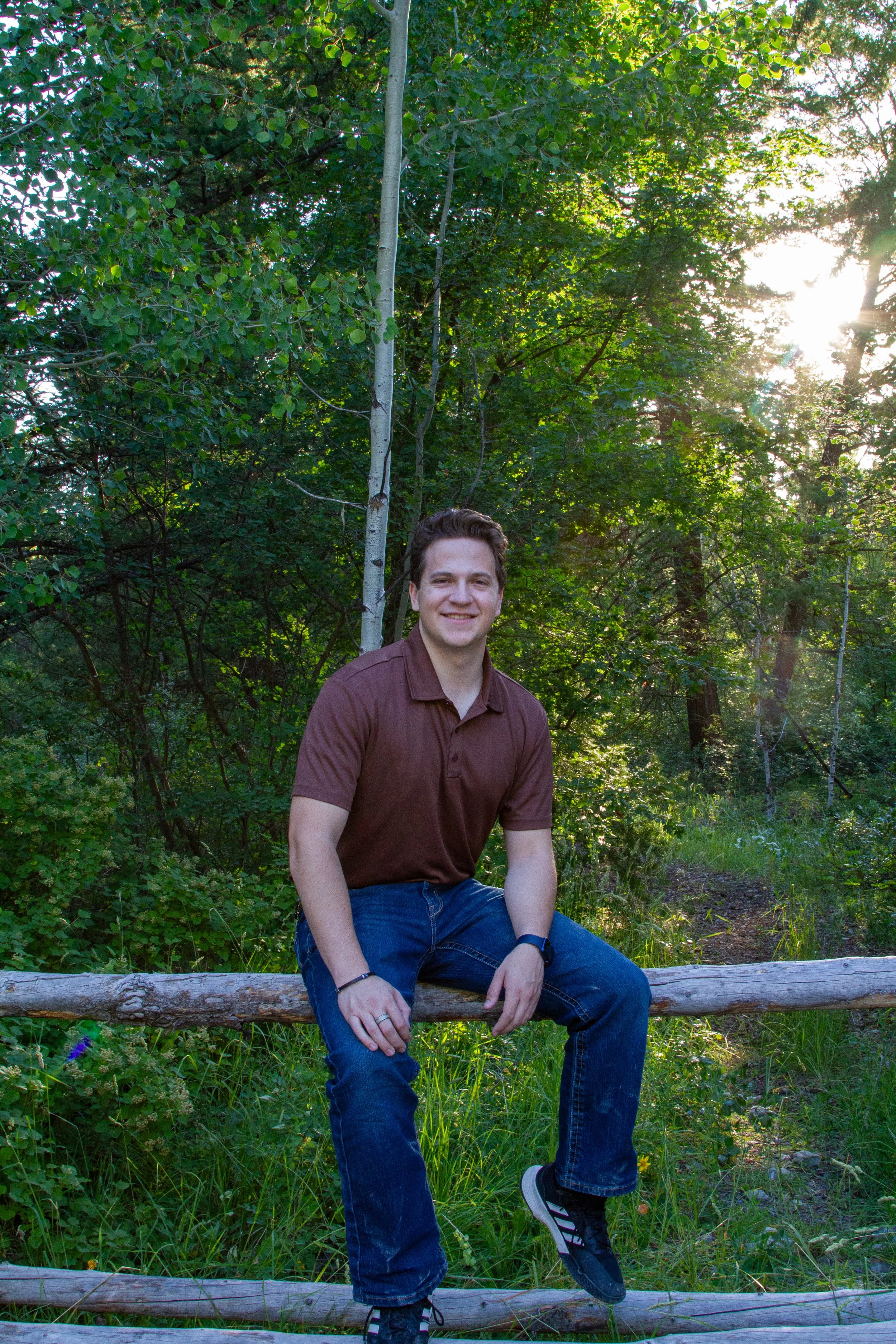 A young man sitting on a log in a natural wooded setting with green leaves and trees, illuminated by sunlight in the background, smiling at the camera.
