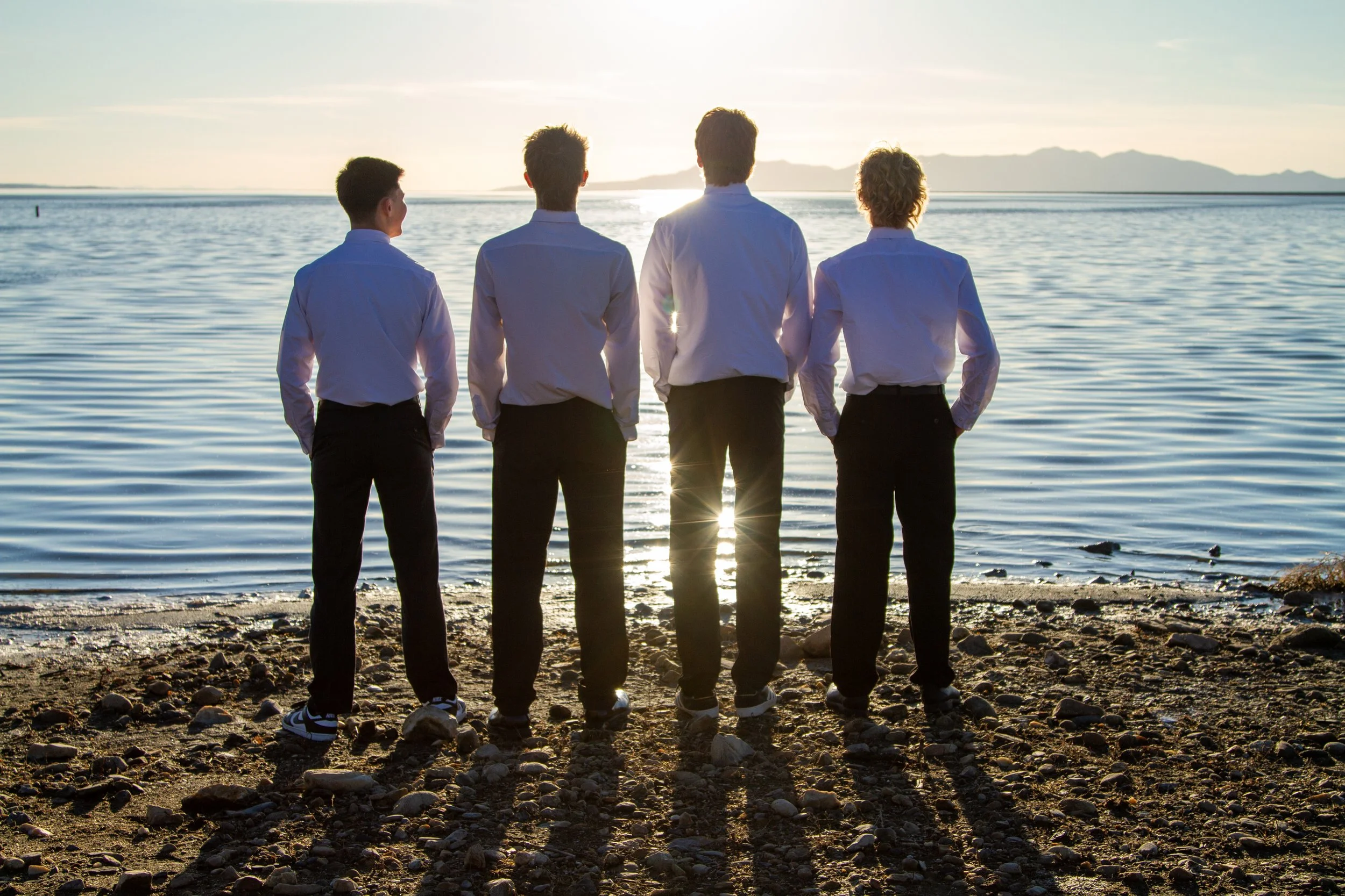 Four young men dressed in white shirts and dark pants standing on a pebbled beach, facing a calm body of water, with mountains in the distance, during sunset.