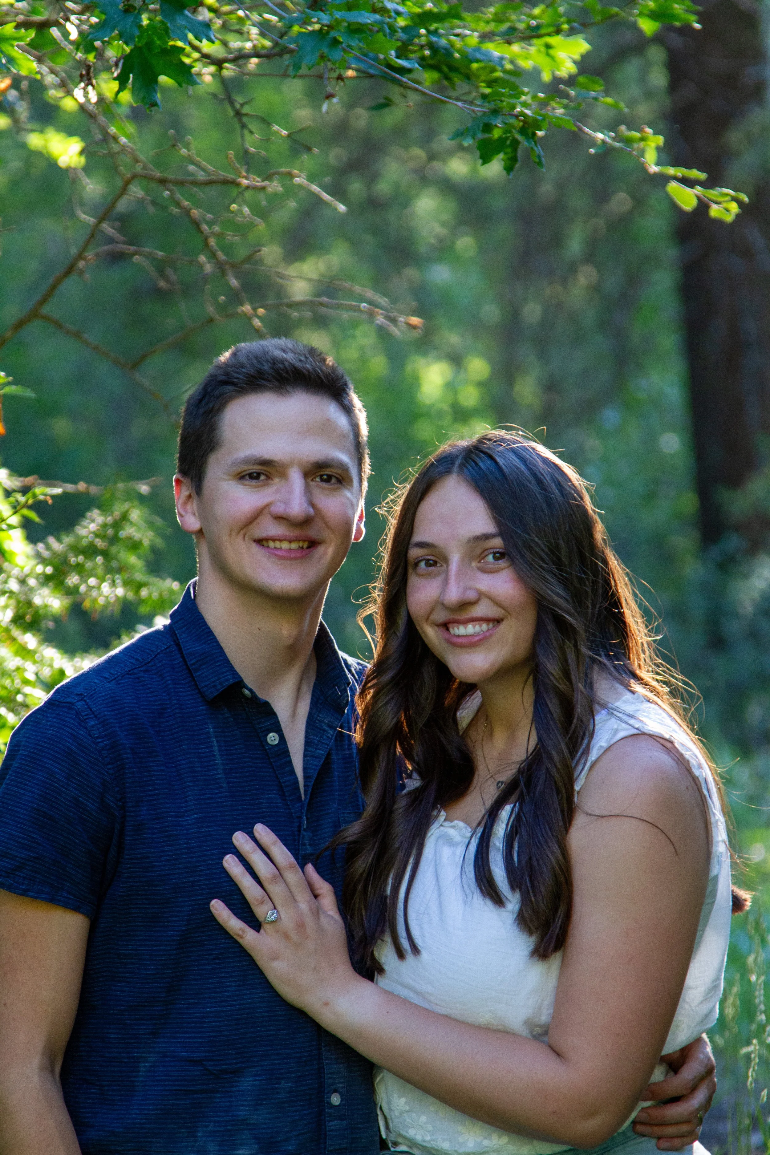 A smiling young man and woman standing close together outdoors in a green, wooded area. The woman has long dark hair and is wearing a white dress, while the man has short dark hair and is dressed in a dark blue shirt. They are embracing in a friendly