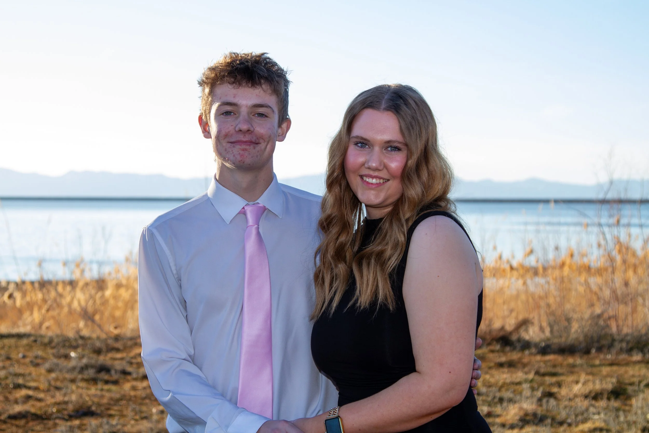 A young man and woman standing outdoors near a body of water, smiling at the camera. The man is wearing a white shirt and pink tie, and the woman is in a sleeveless black dress with wavy brown hair.