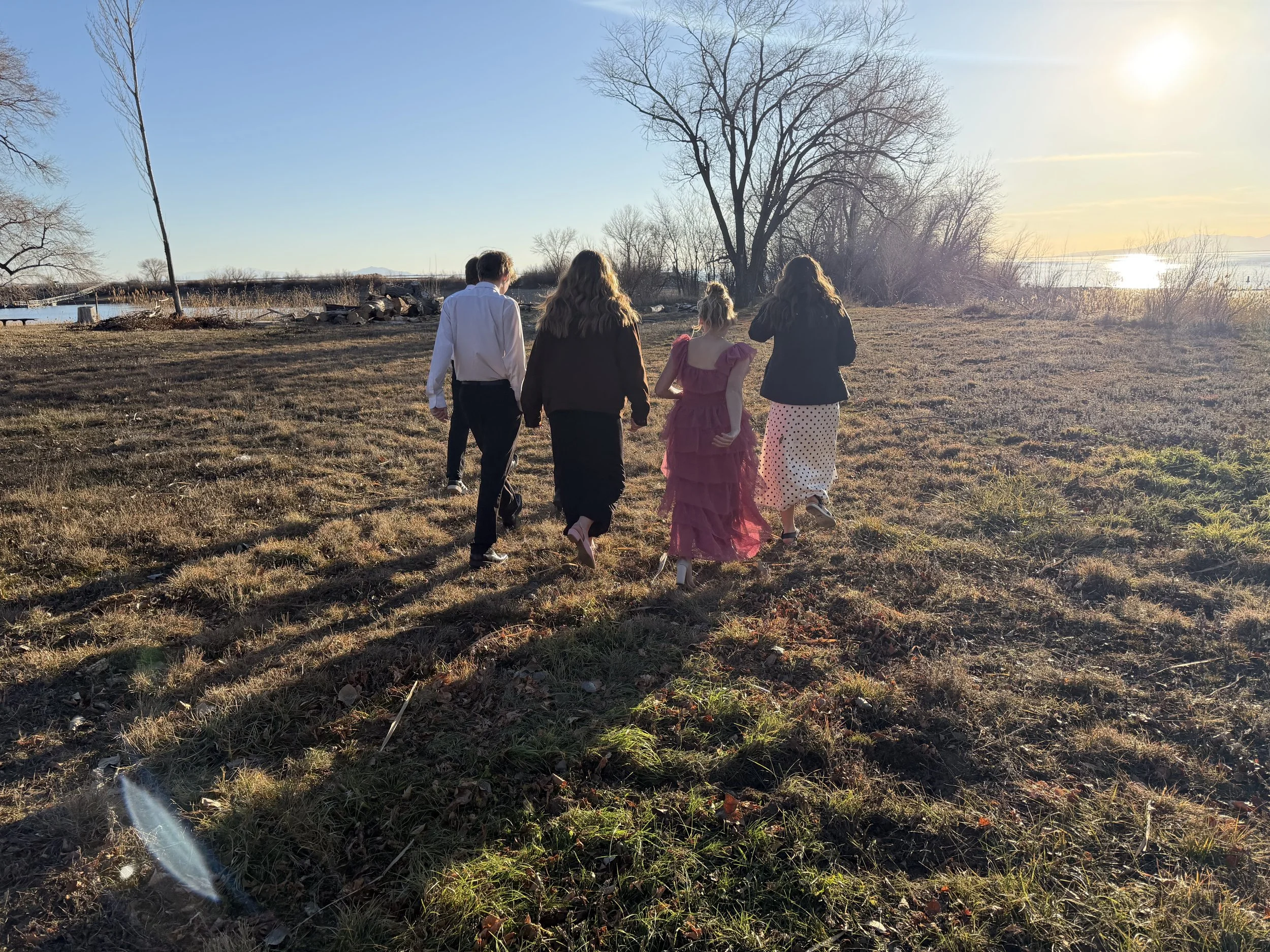 A group of five people walking on a grassy field near a body of water during sunset. The group includes three women and two men, one woman in a pink dress, and the others dressed in dark and light trousers and jackets. Bare trees are visible in the b