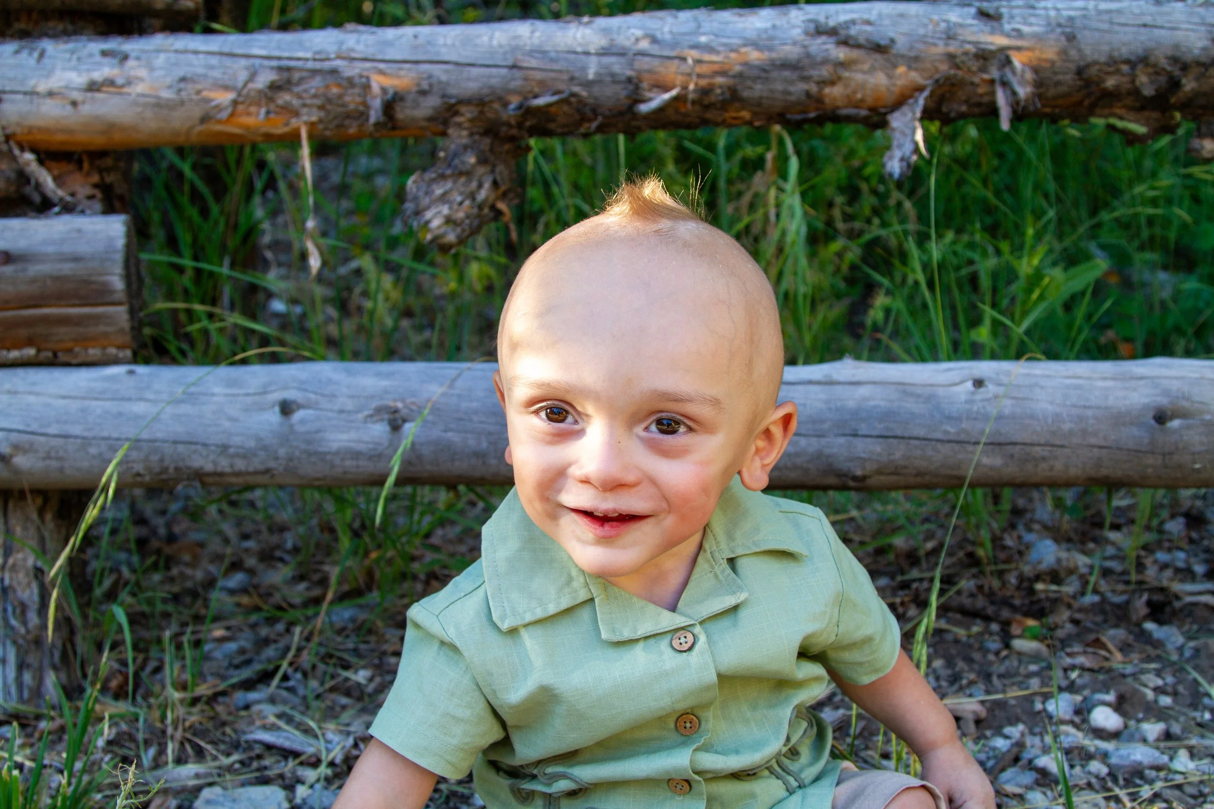 A young boy with a light-colored, short hairstyle and big brown eyes, smiling and leaning back on the ground in front of a rustic wooden fence with green grass behind it.