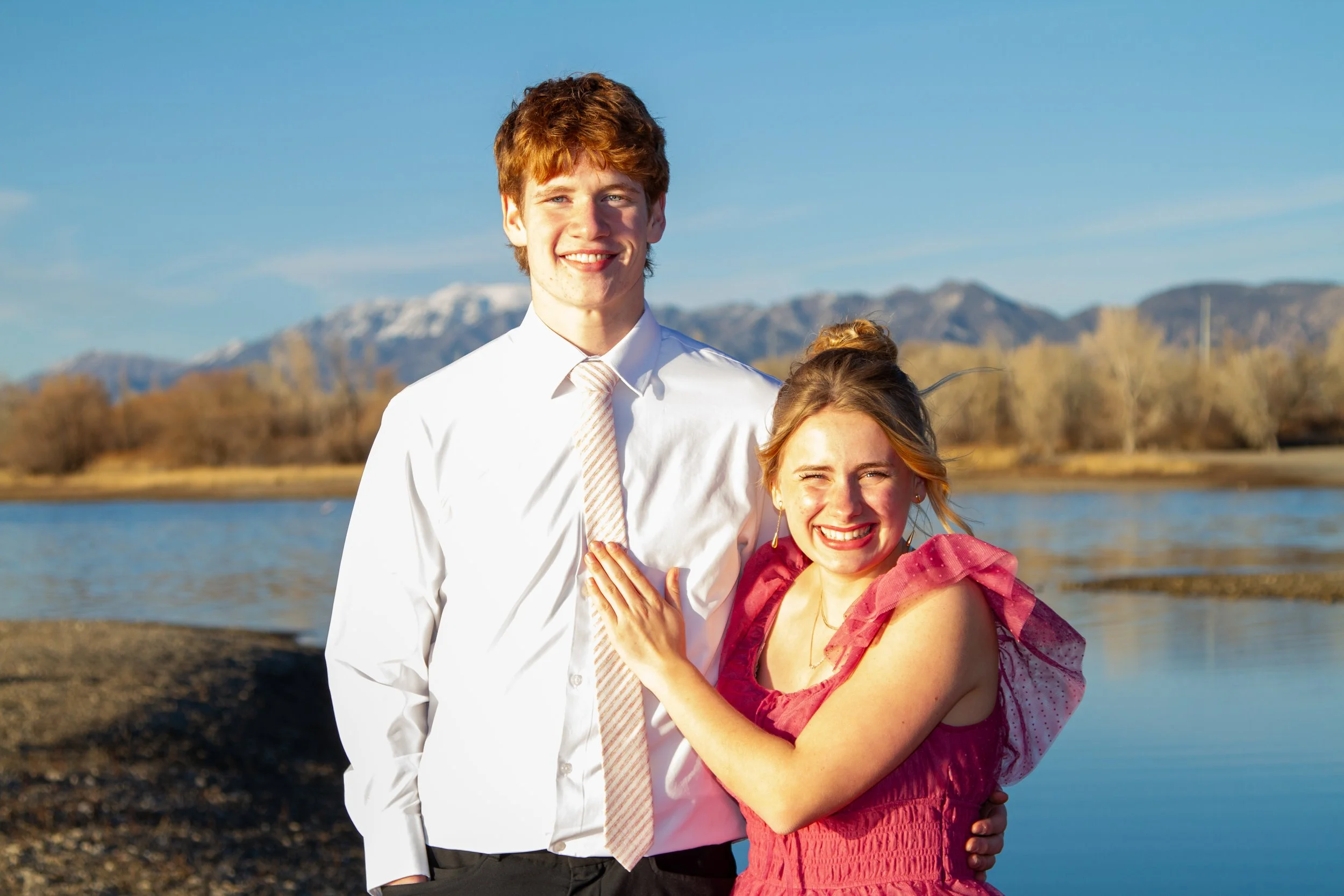 A smiling young man in a white shirt and striped tie standing next to a woman in a red dress with layered ruffles on her shoulders, near a body of water with mountains in the background on a sunny day.