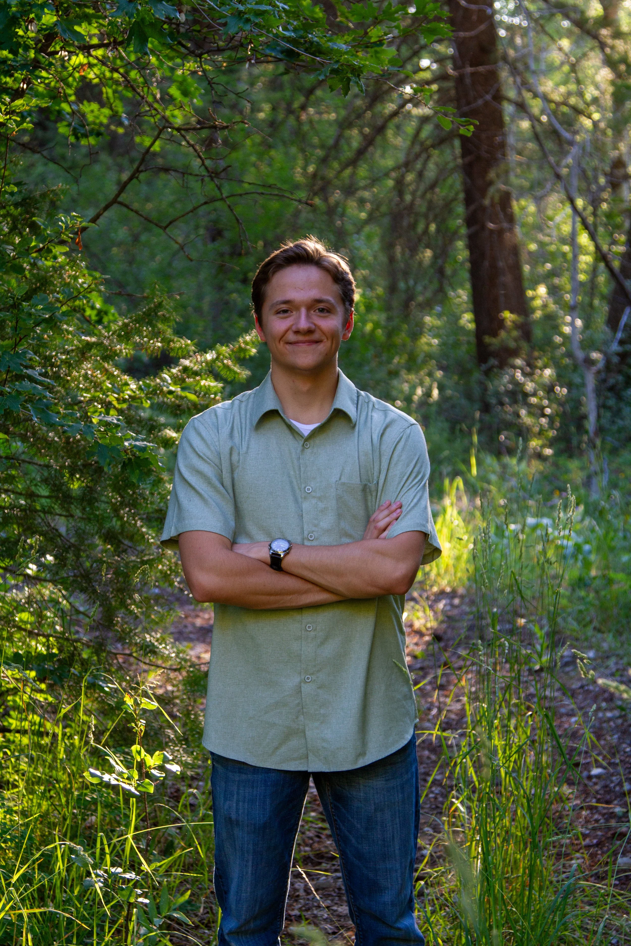 A young man with brown hair smiling in a green forest, crossing his arms, wearing a light green shirt and jeans, with sunlight filtering through the trees.