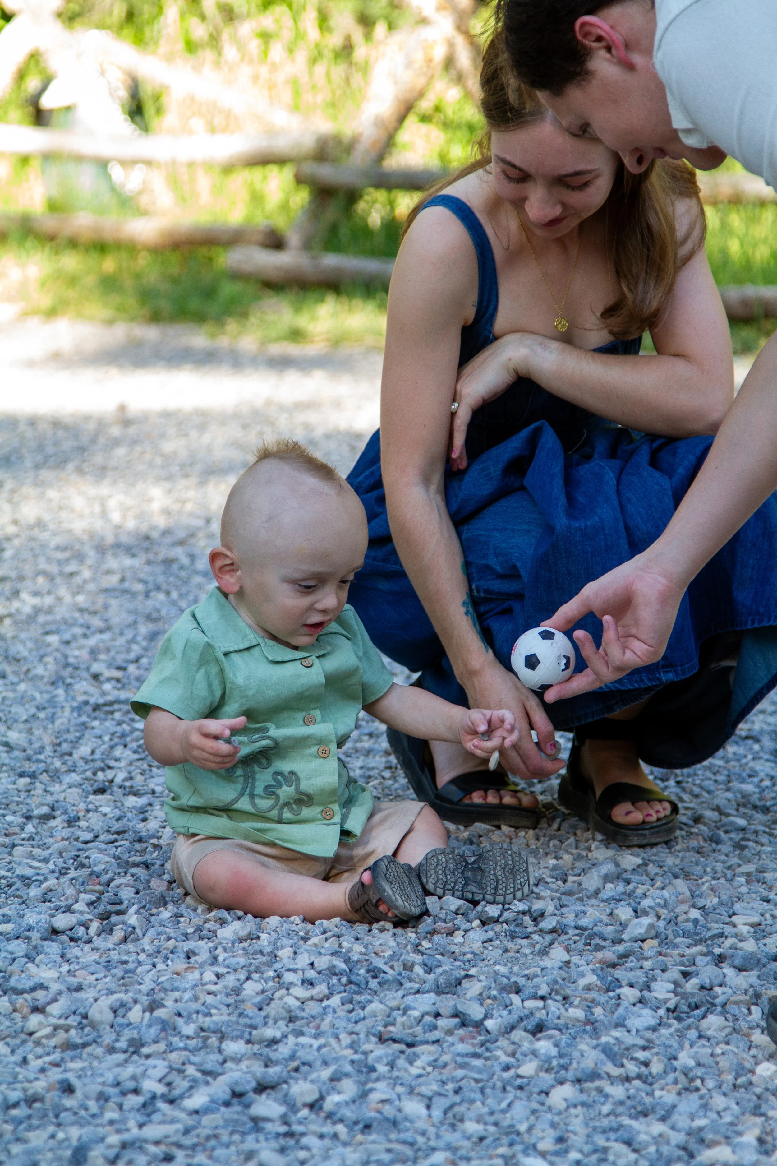 A group of children and a woman are outdoors on a gravel surface. The woman and an older child are handing a small soccer ball to a young child sitting on the ground. The scene is bright with greenery in the background.