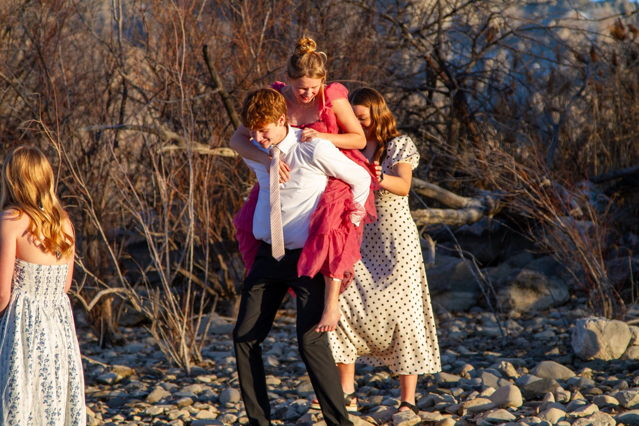A group of young women at a rocky outdoor location with leafless trees, one of whom is giving a piggyback ride to another in a pink dress while the others watch and smile.