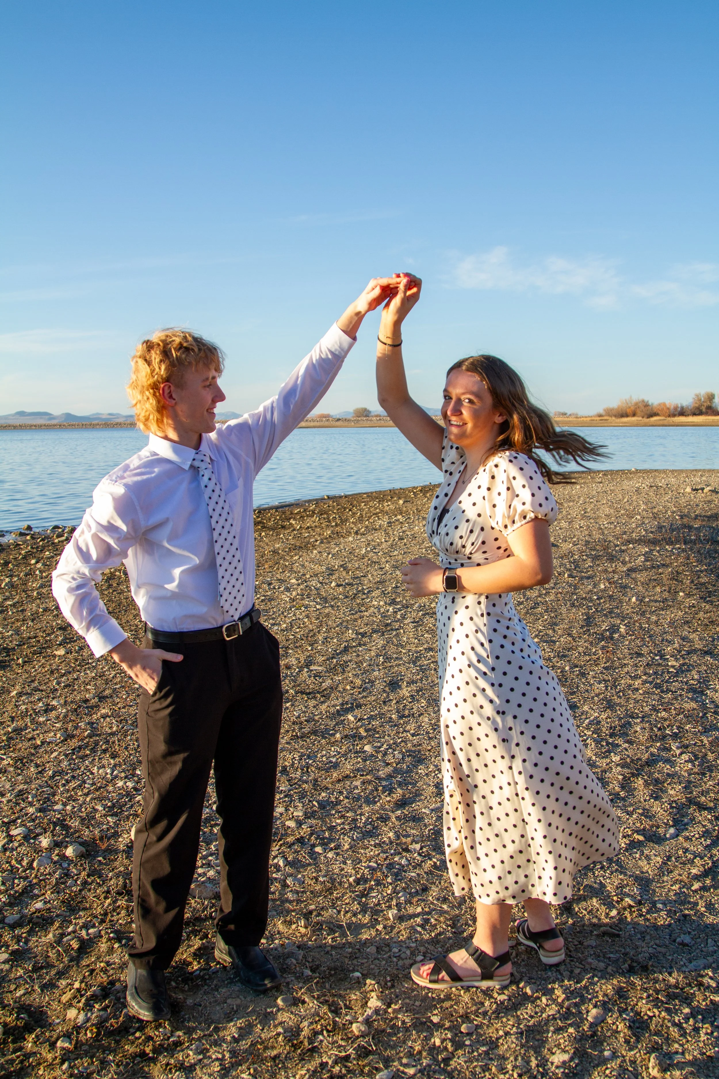 A couple on a rocky shoreline by a lake, celebrating with a high-five in sunny weather.