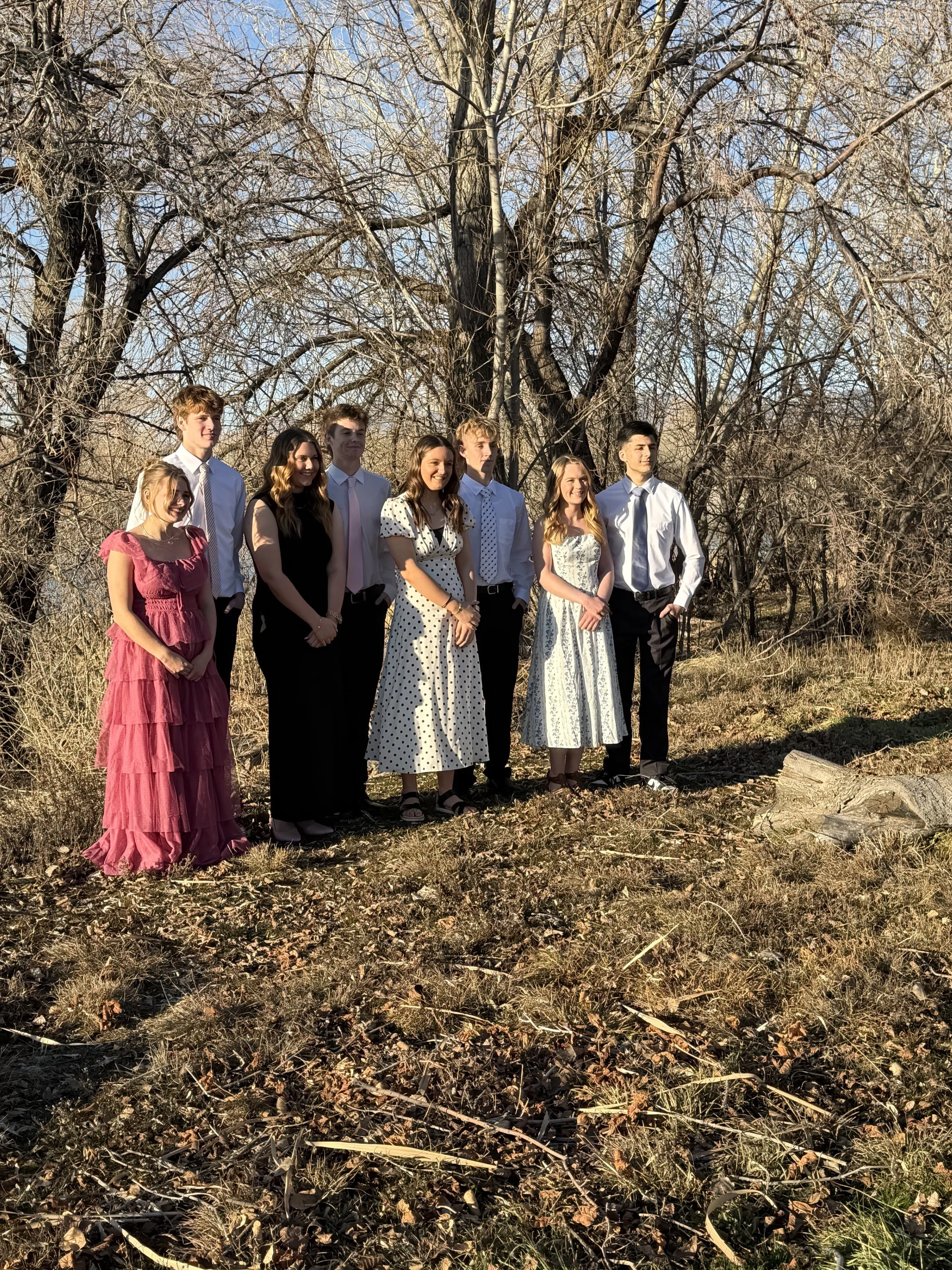 Group of eight teenagers dressed in formal attire outdoors on a sunny day with leafless trees in the background.