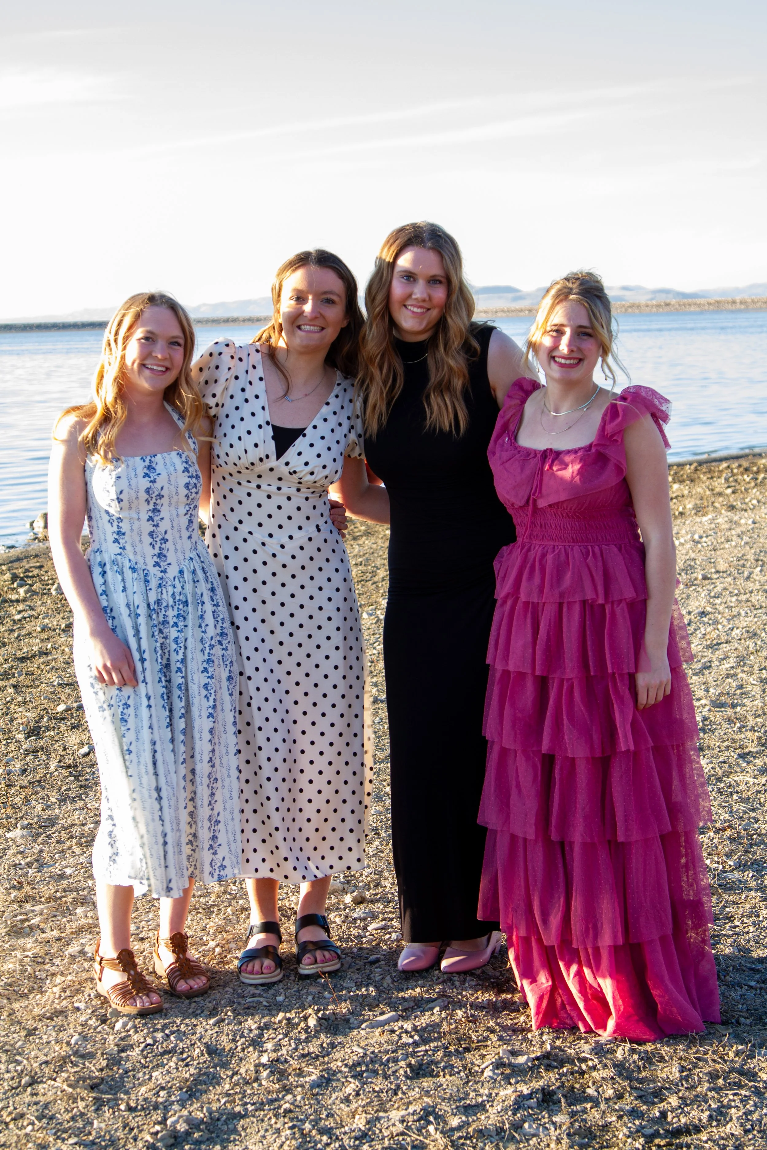 Four women in dresses standing on a rocky beach near a body of water, smiling at the camera during sunset or late afternoon.