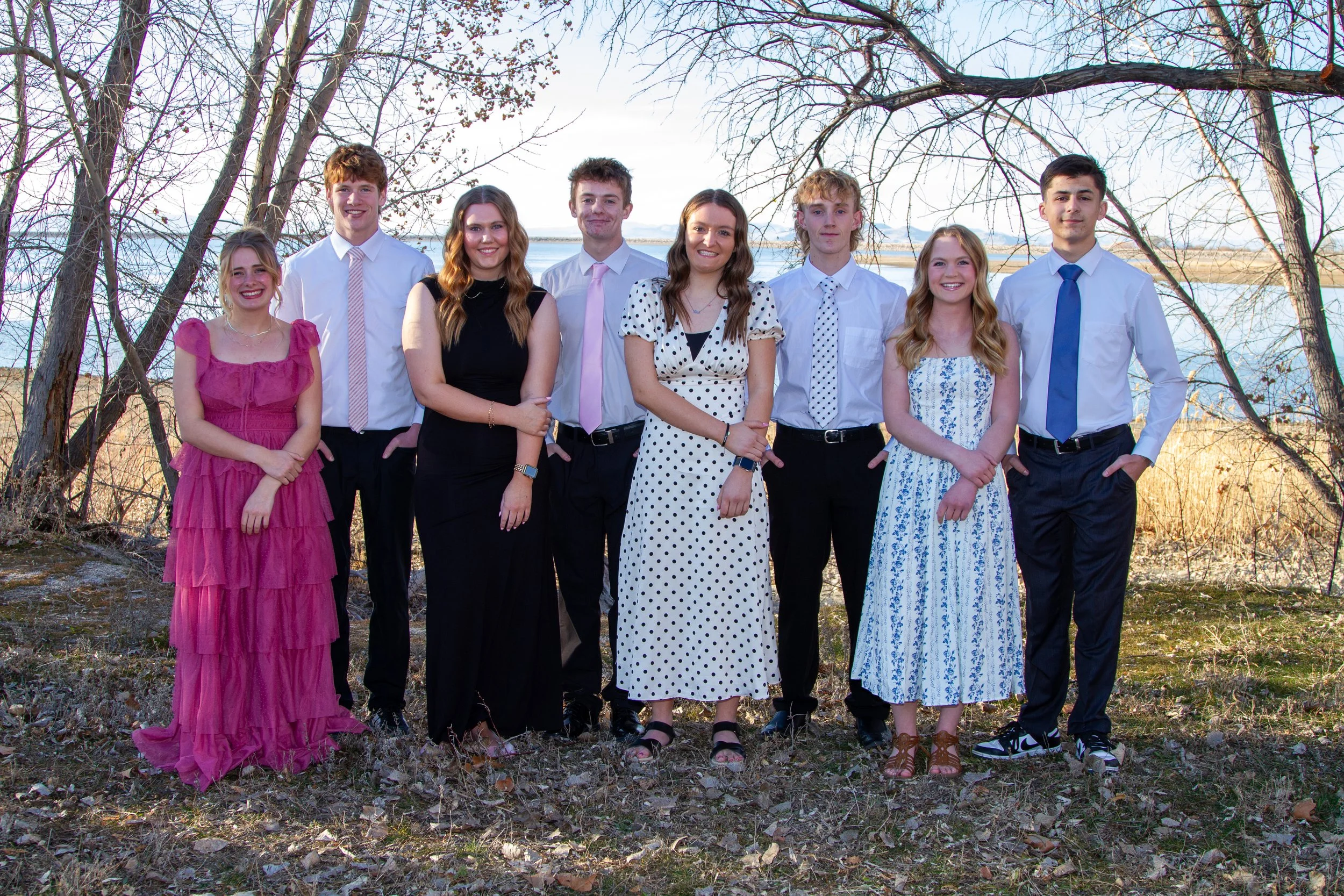 Group of nine young adults standing outdoors on a grassy area near a body of water with trees in the background, dressed in semi-formal attire.