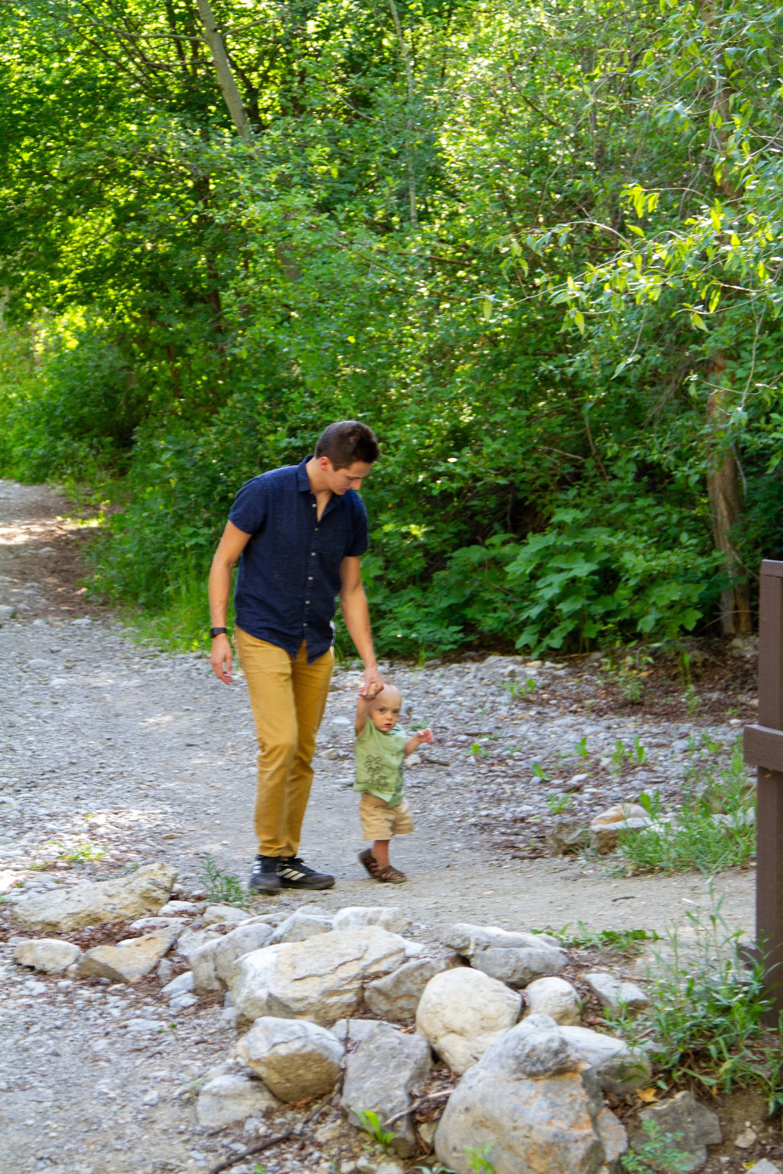 A man and a young girl walking along a gravel and rocky trail in a wooded area, surrounded by green foliage.