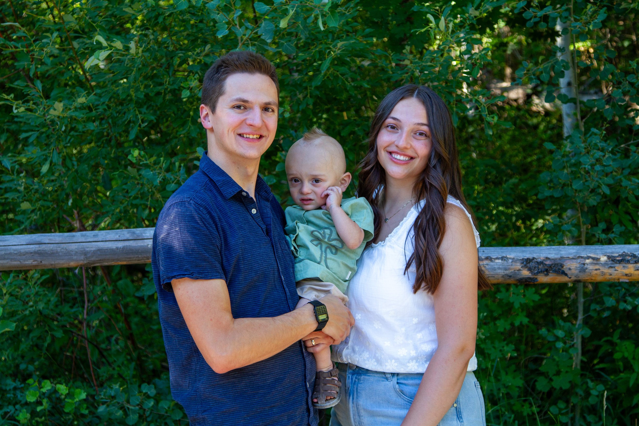 A family of three, a man, woman, and young child, standing outdoors in front of green foliage, smiling at the camera. The man is wearing a blue shirt, the woman a white sleeveless top, and the child is dressed in green.
