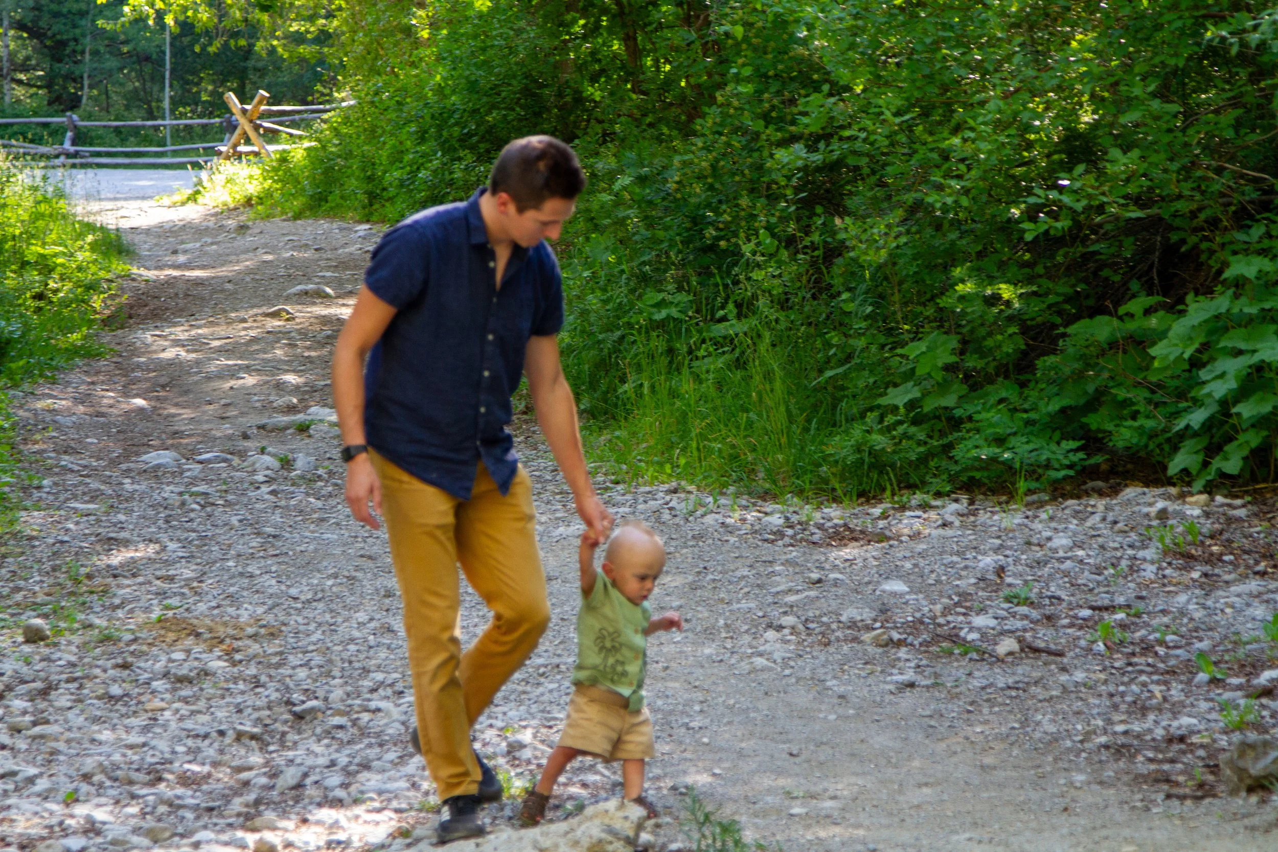 A man and a young child walking hand in hand on a gravel trail in a lush green forest, with sunlight filtering through the trees.