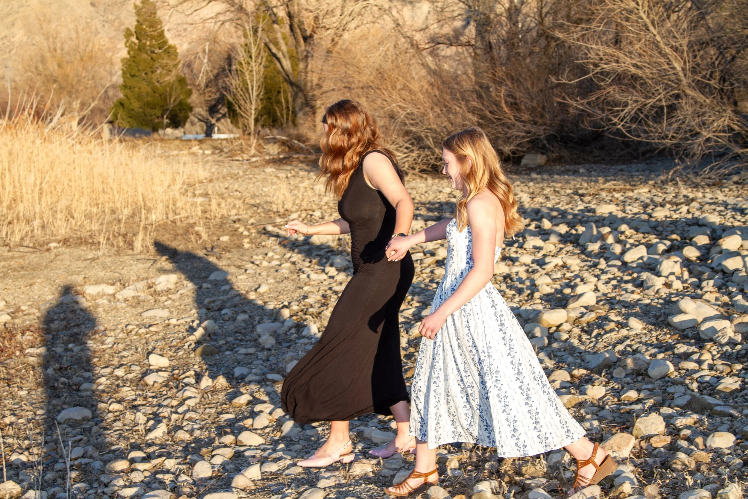 Two young women with red hair walking on a rocky beach, holding hands, during sunset.