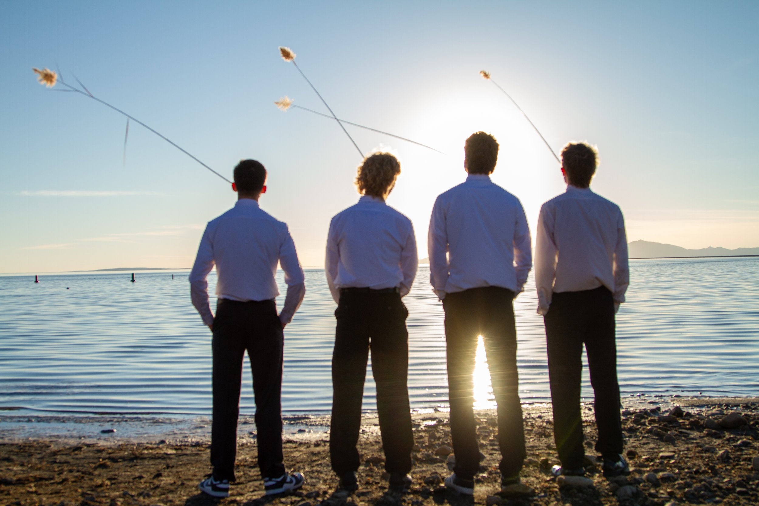 Four young men in white shirts and black pants standing on a rocky beach at sunset, facing the water, four kites flying in the sky.