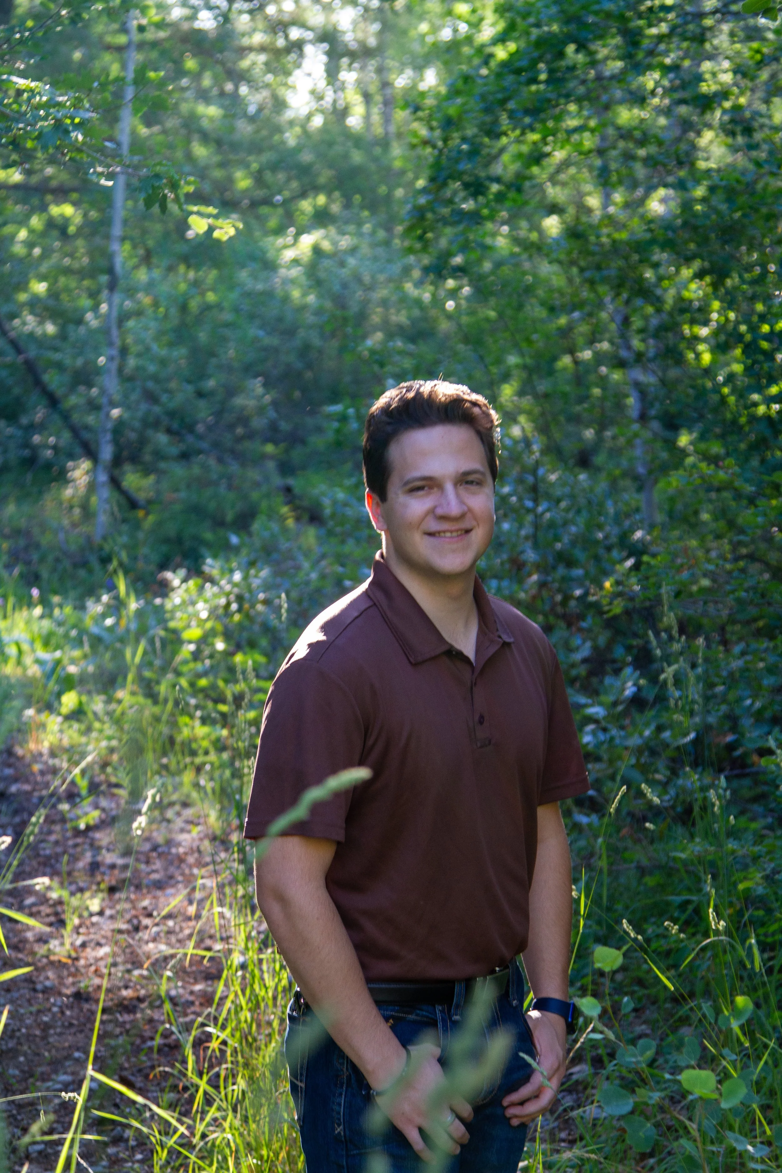 A young man with dark hair smiling, wearing a brown polo shirt and jeans, standing outdoors in a green, wooded area with sunlight filtering through the trees.