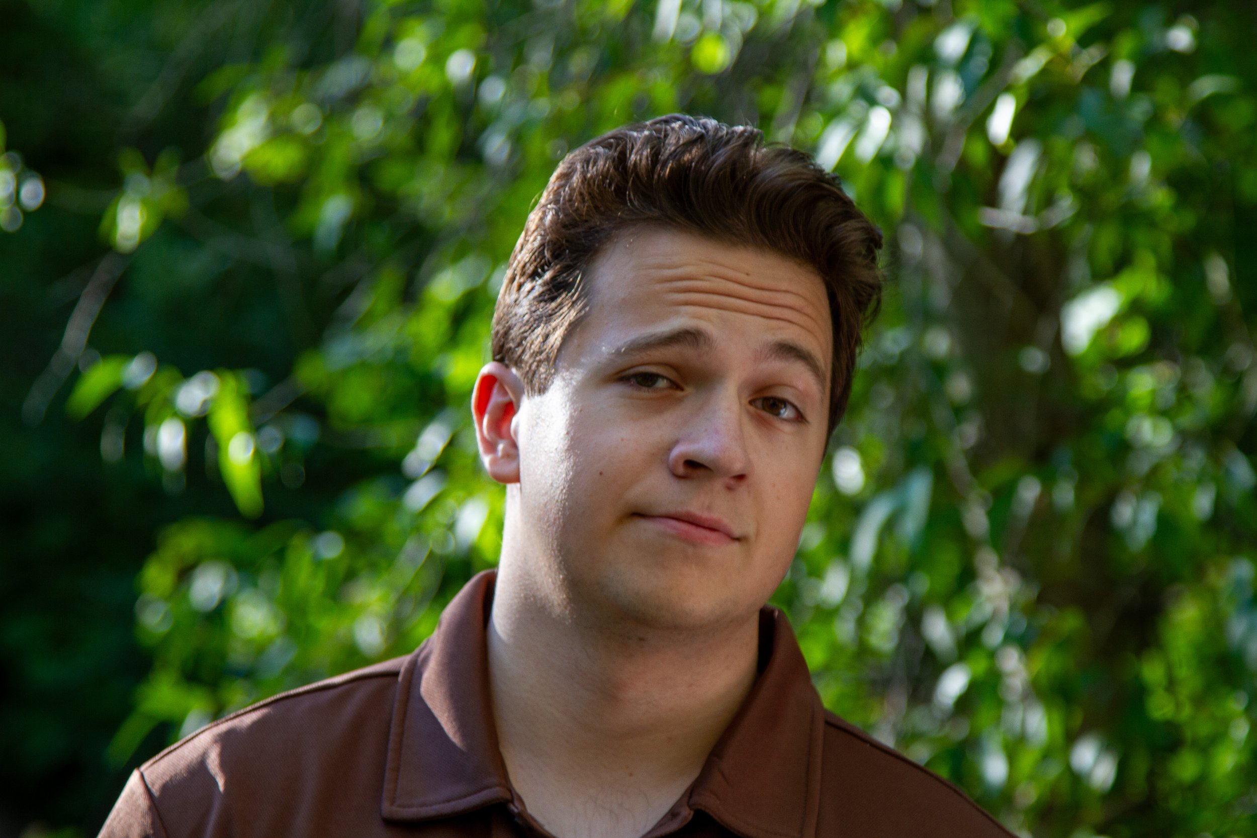 A young man with brown hair, wearing a brown jacket, looking slightly to the side with a subtle smile, standing outdoors with green leafy trees in the background.