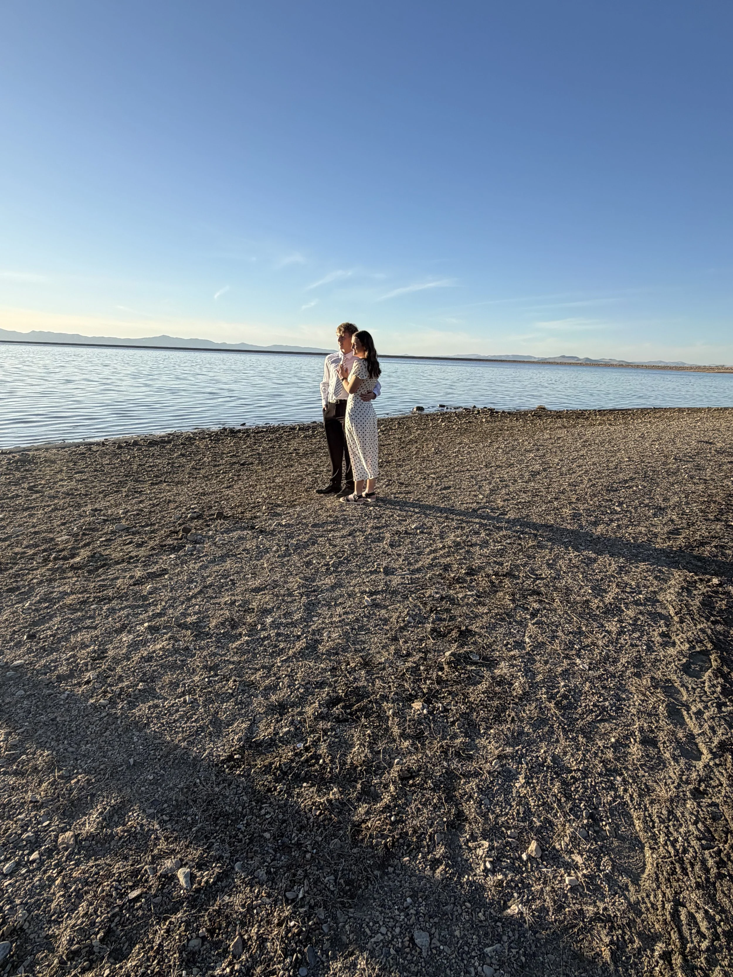 A couple standing arm in arm on a sandy beach next to a body of water, with mountains in the distance and a clear blue sky.