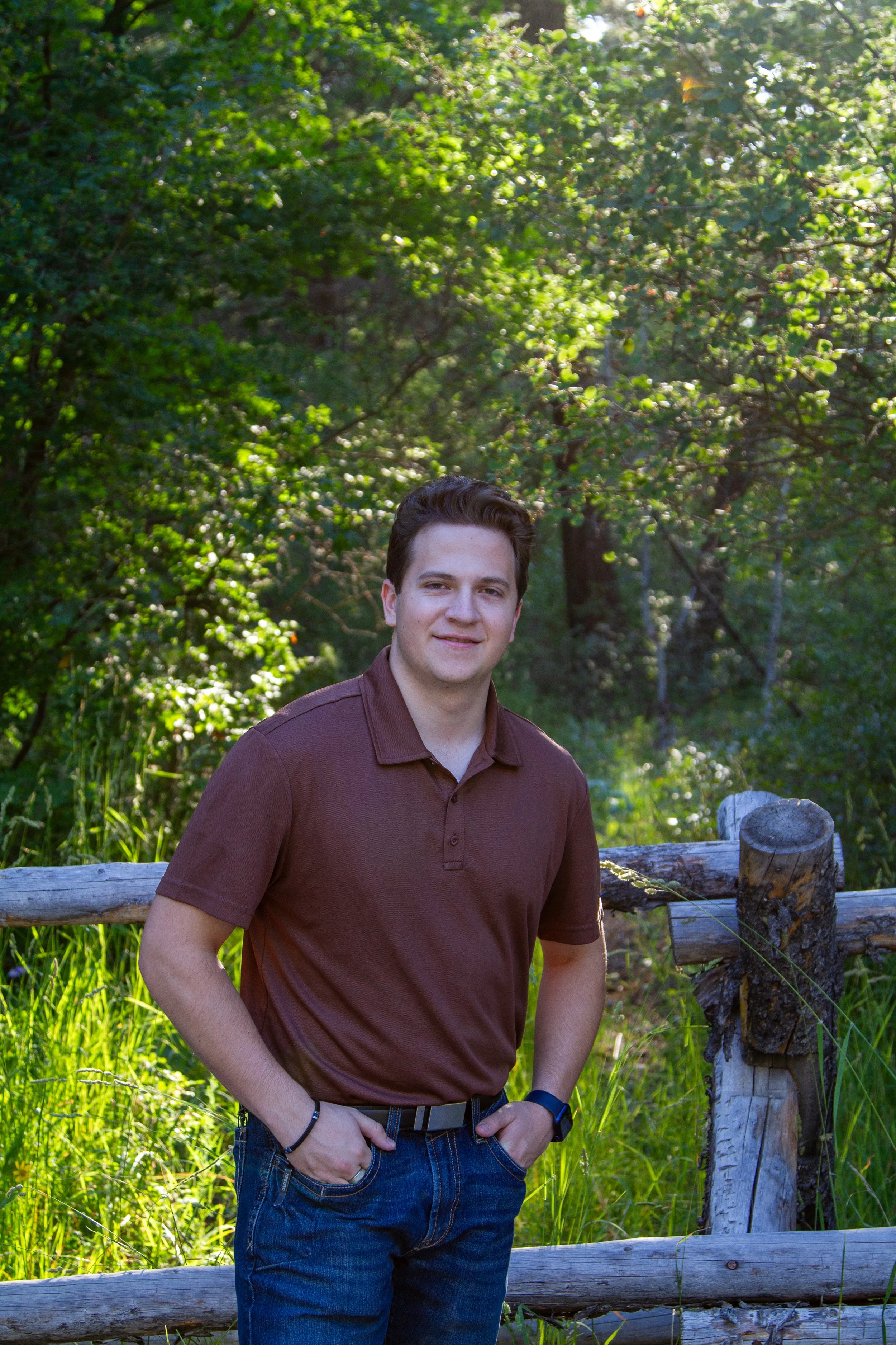 A young man with brown hair smiling and standing outdoors in front of a wooden fence, with green trees and bushes in the background.