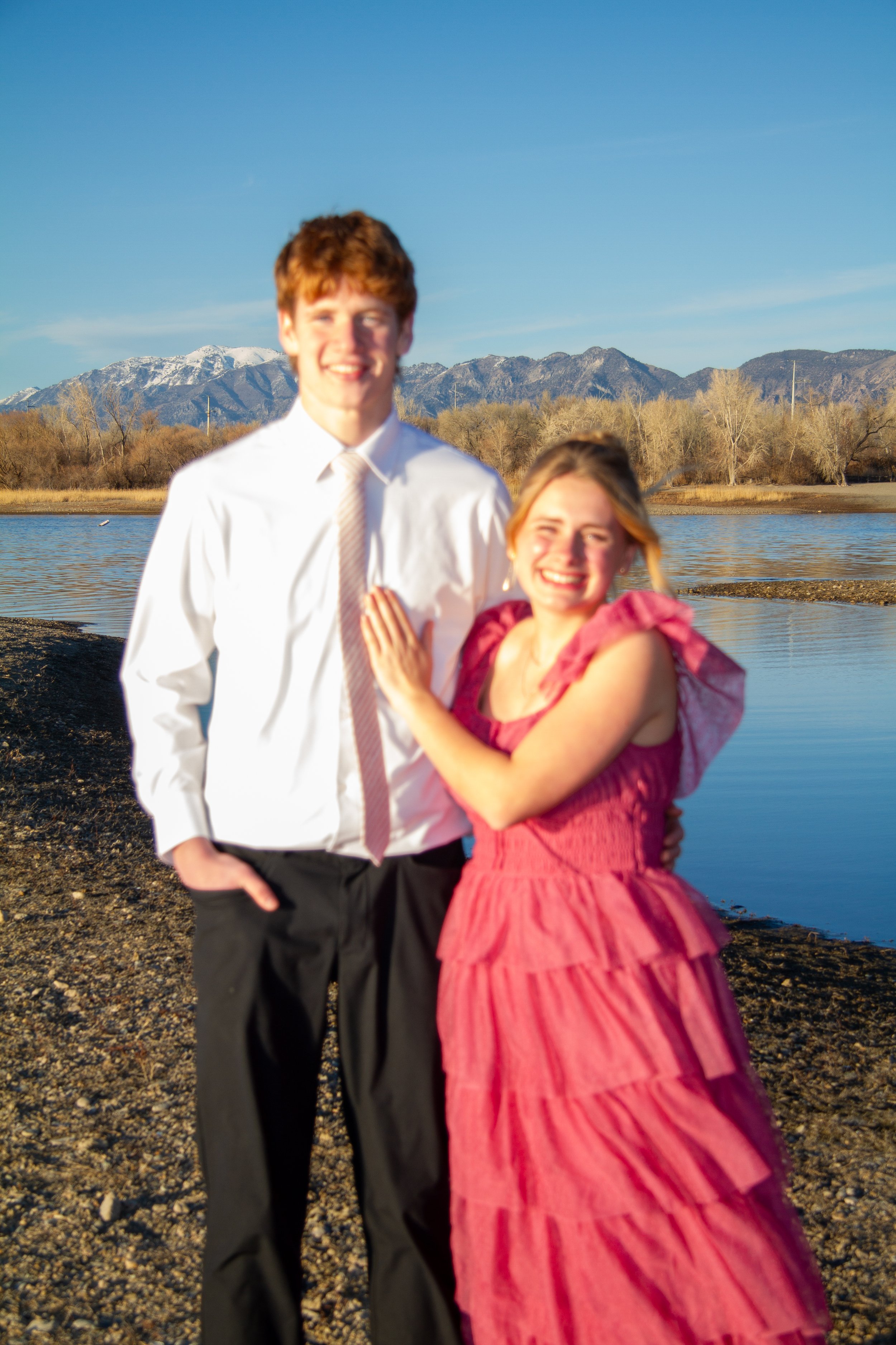 A smiling young man and woman standing by a river with mountains in the background, during daytime.