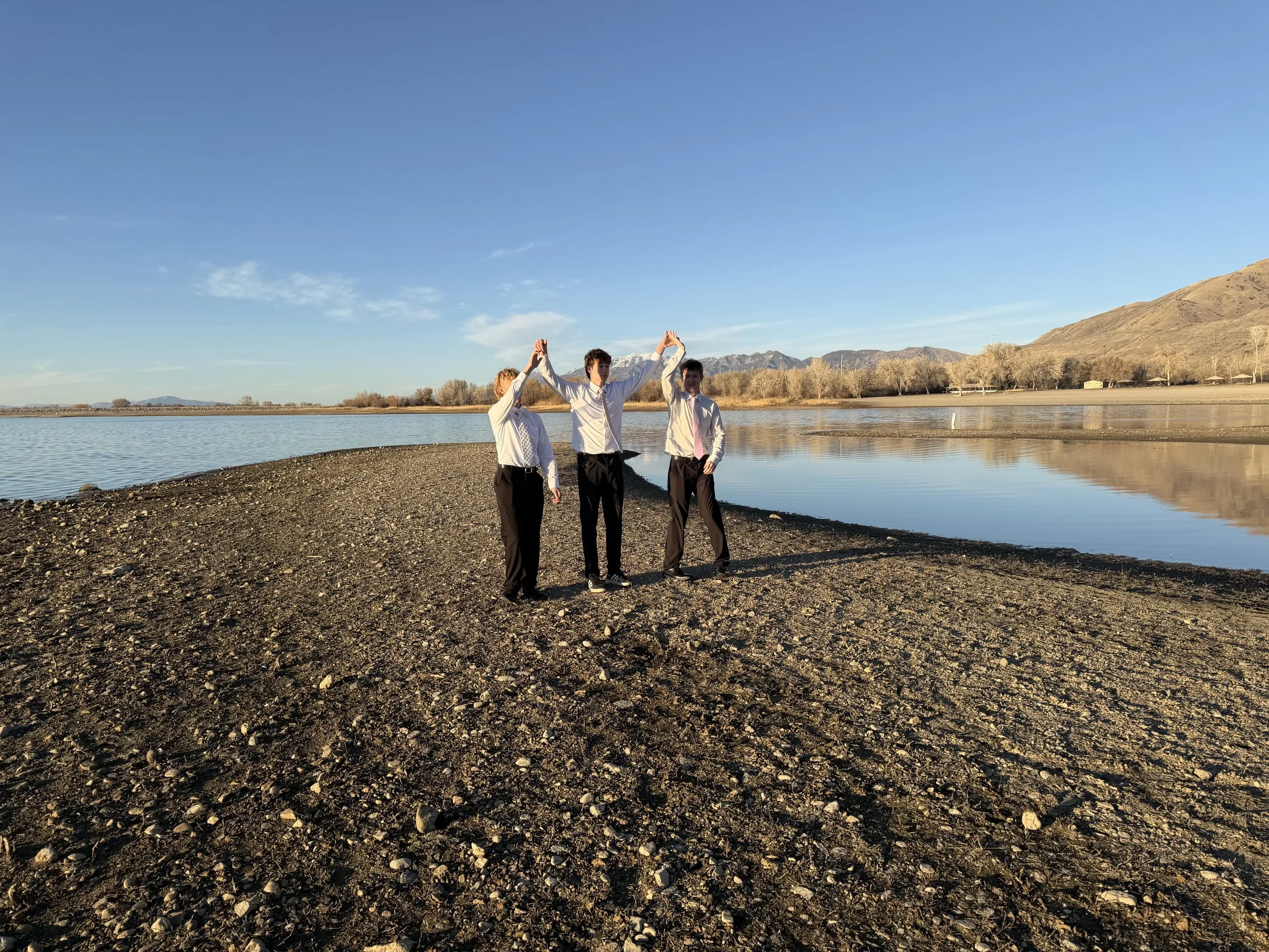 Three men in white shirts and dark pants are on a rocky shoreline near a calm body of water, with mountains and trees in the background, during sunset or sunrise.