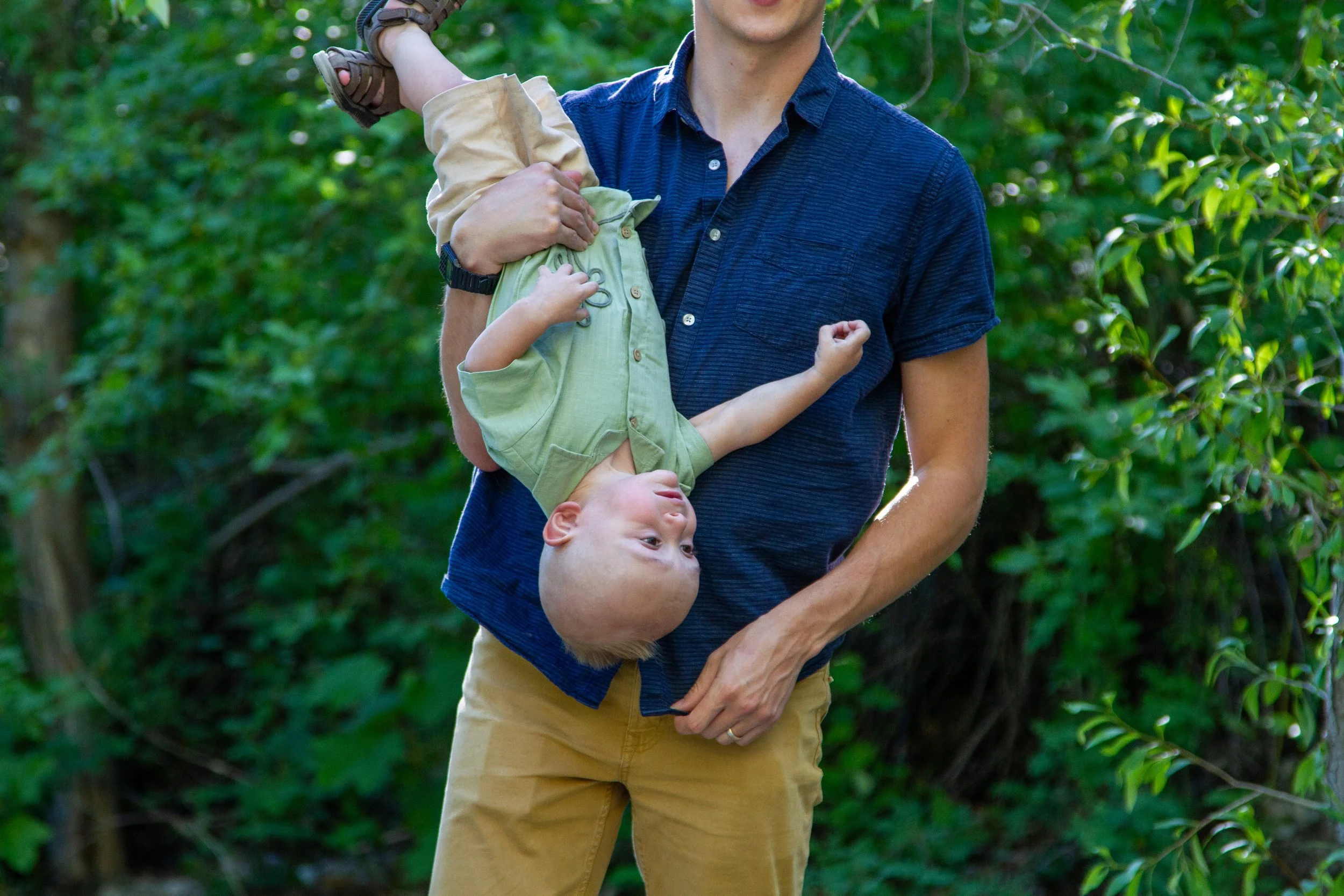 A man holding a young child upside down in a forest with green trees and sunlight.