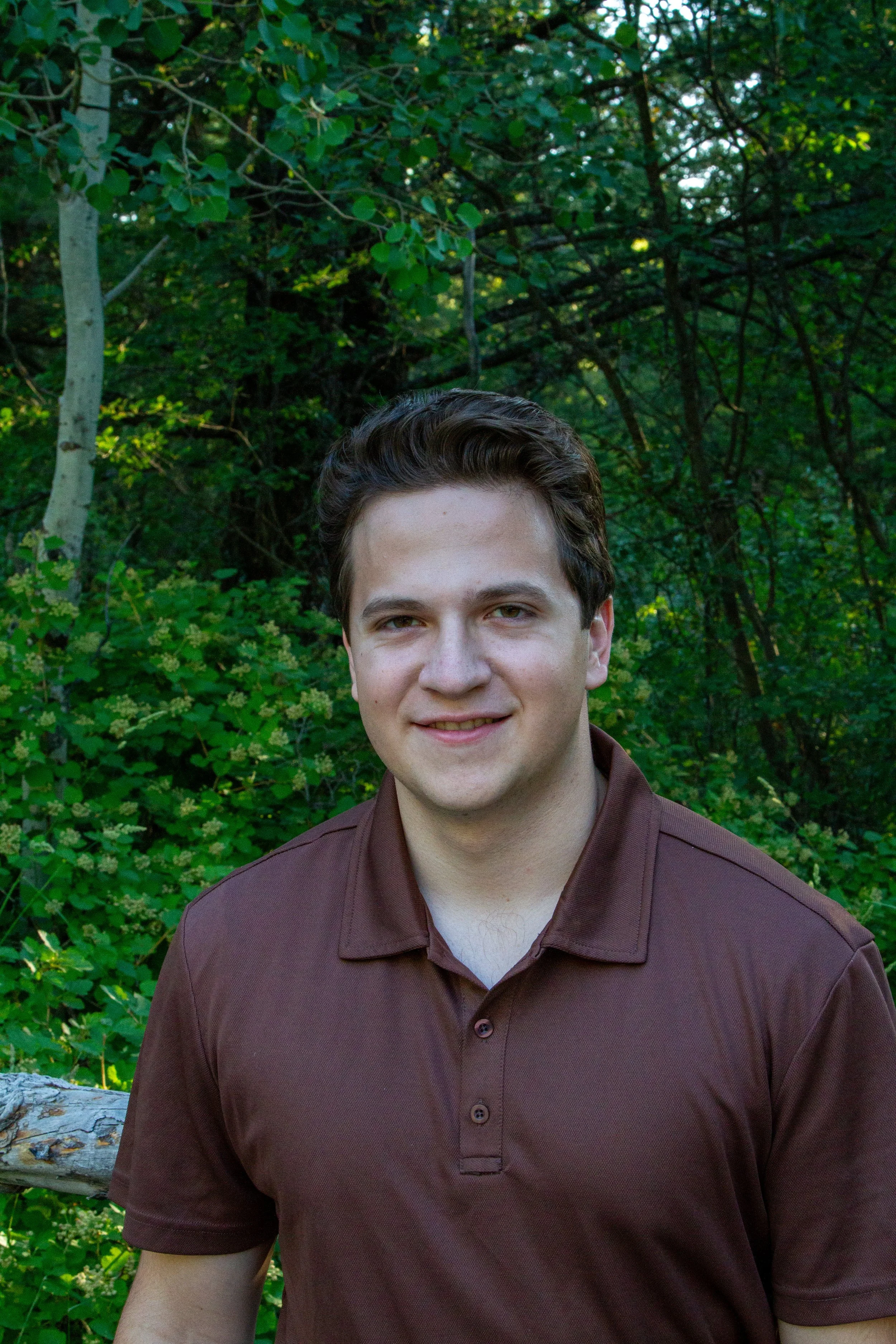 Close-up of a young man with brown hair smiling, wearing a brown button-up shirt, outdoors among green trees and foliage.