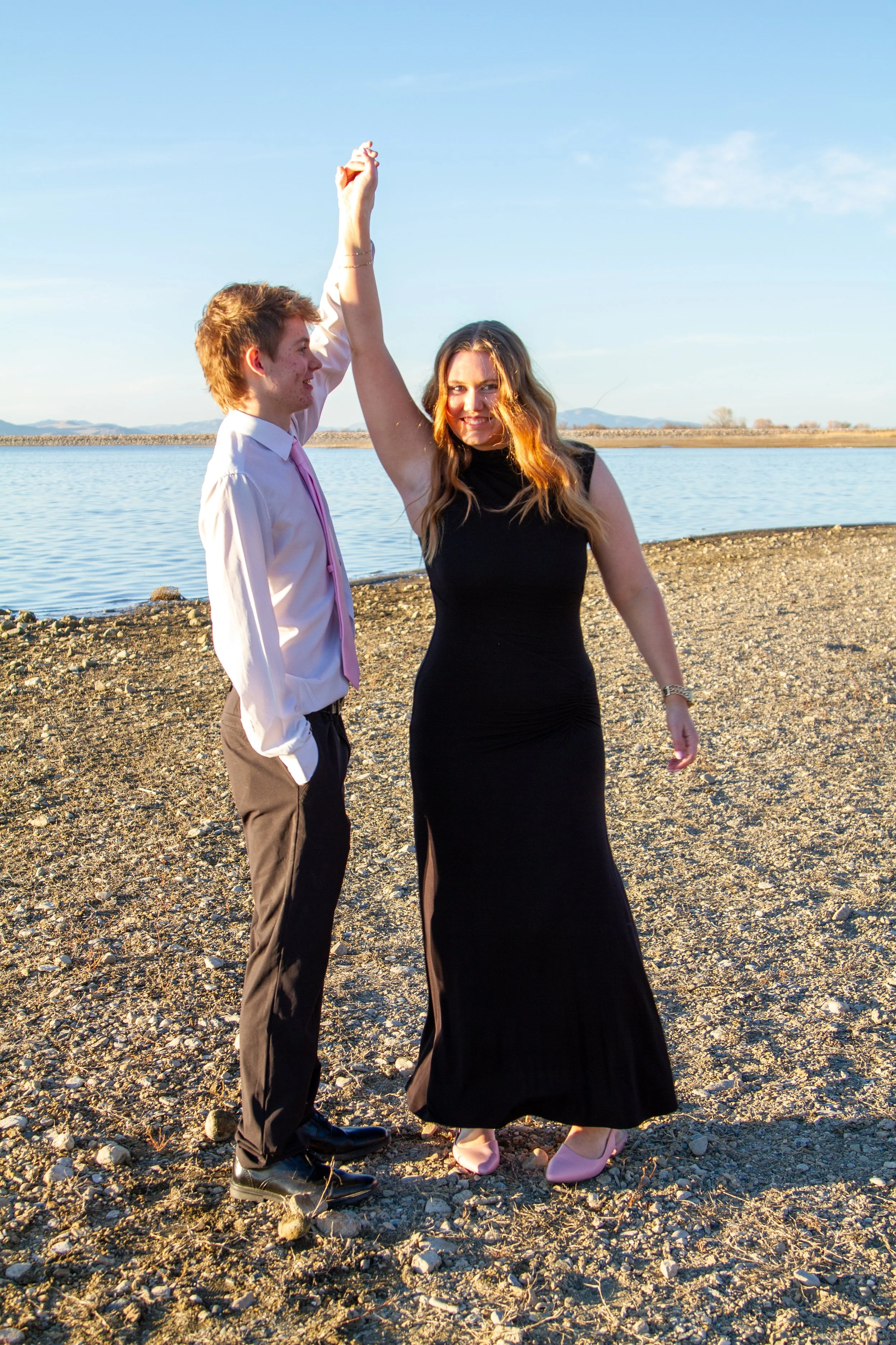 A young man and woman standing on a rocky shoreline near a body of water, holding hands with the woman's arm raised high, with a clear blue sky in the background.