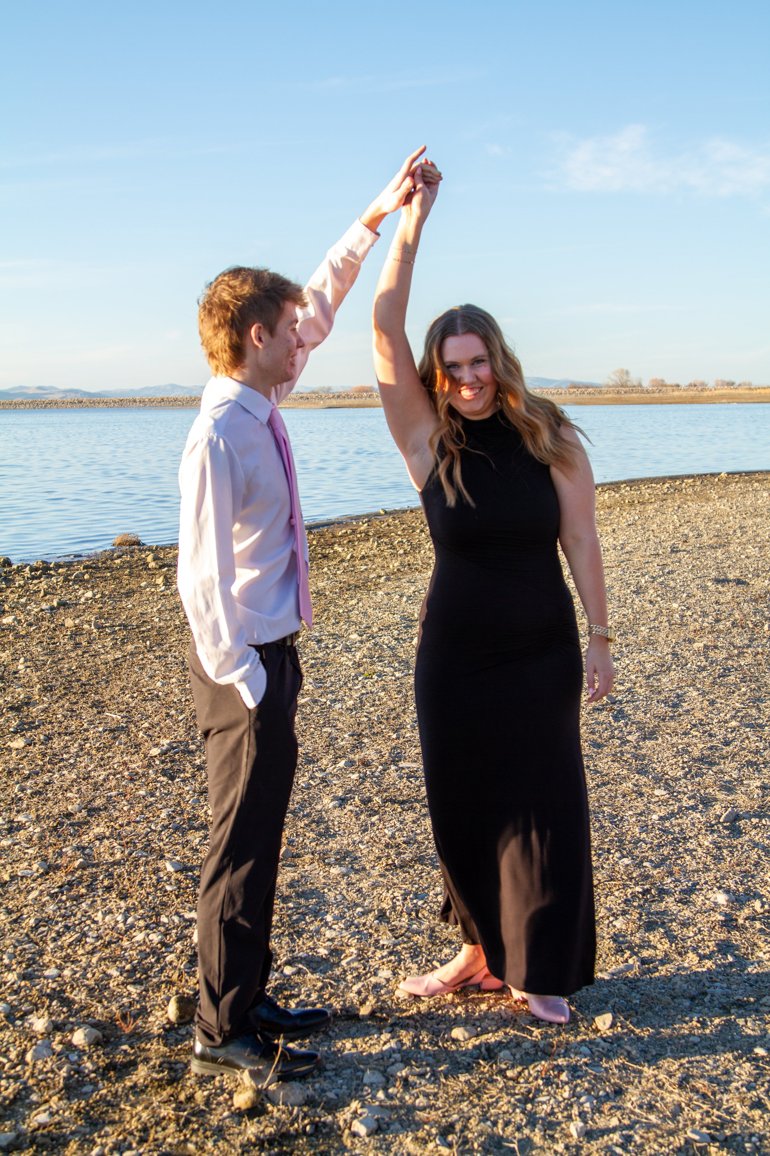 A young man in a white shirt and gray pants dances with a woman in a black dress on a rocky beach by the water, holding hands and smiling.