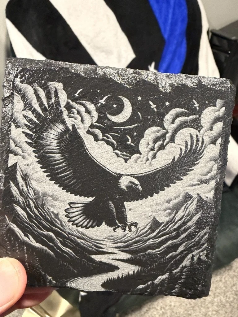 A black and white artwork depicting an eagle soaring over mountains with a cloudy sky, a crescent moon, and stars.