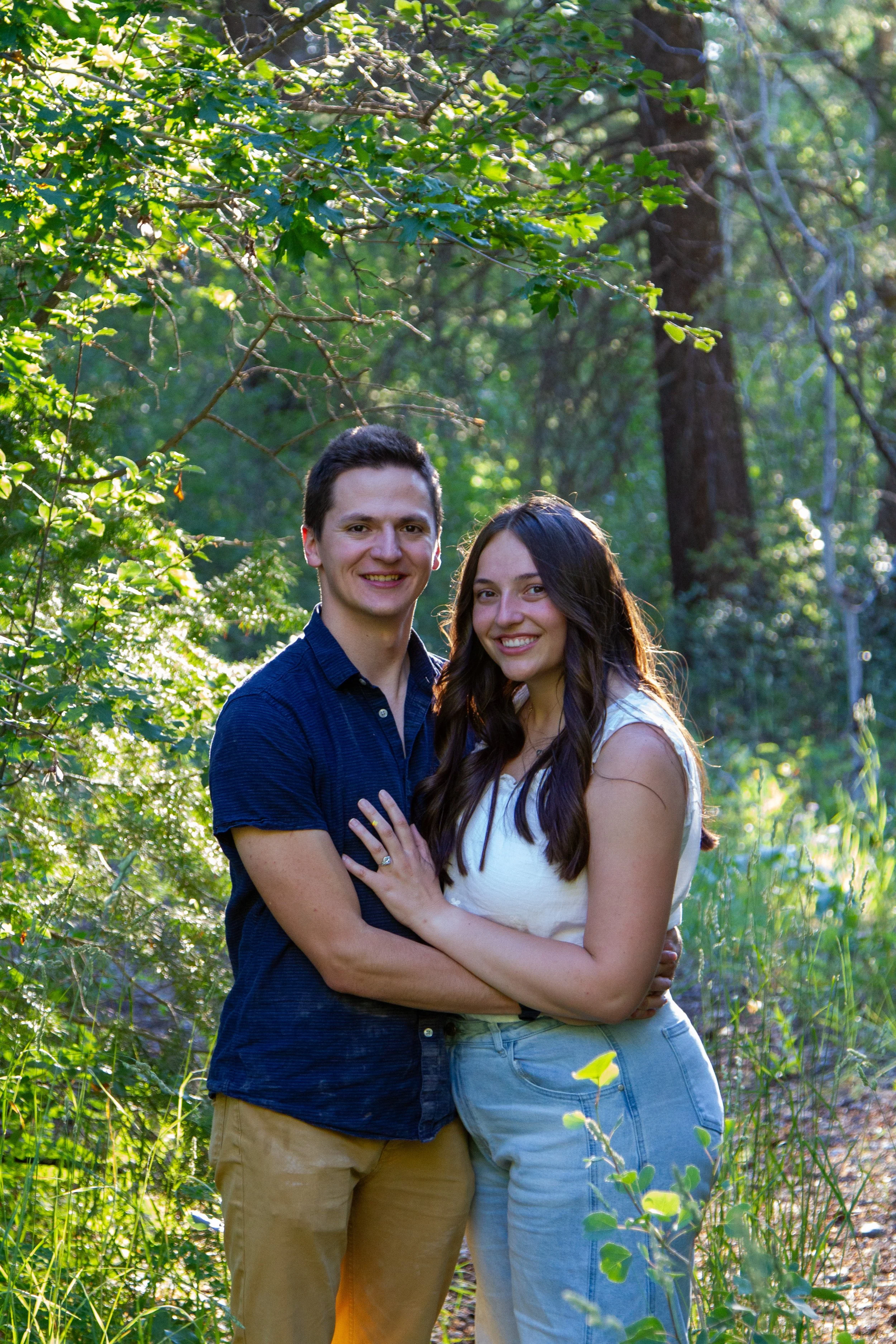 A young couple smiling and hugging in a lush green forest during daytime.