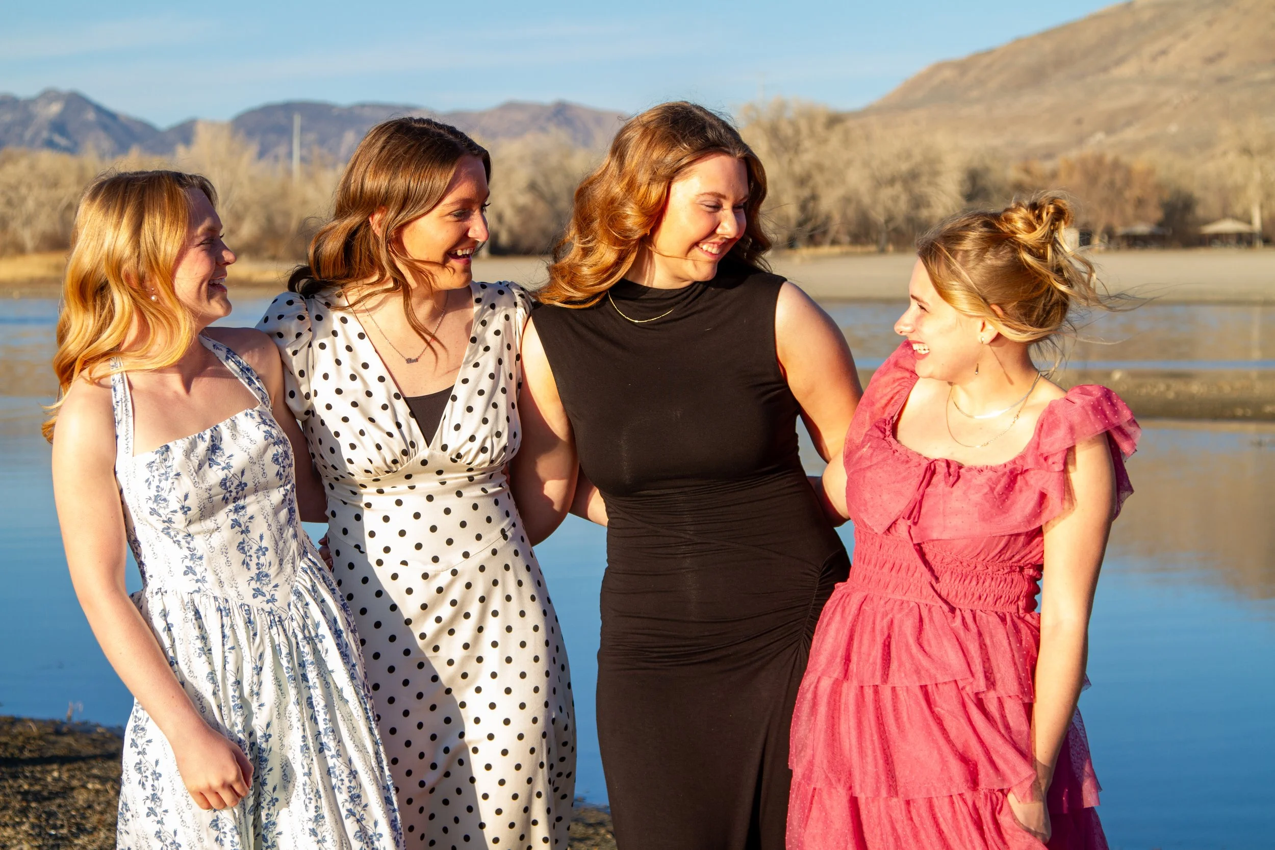 Four women standing on a shoreline near a lake, smiling and talking to each other, with mountains in the background.