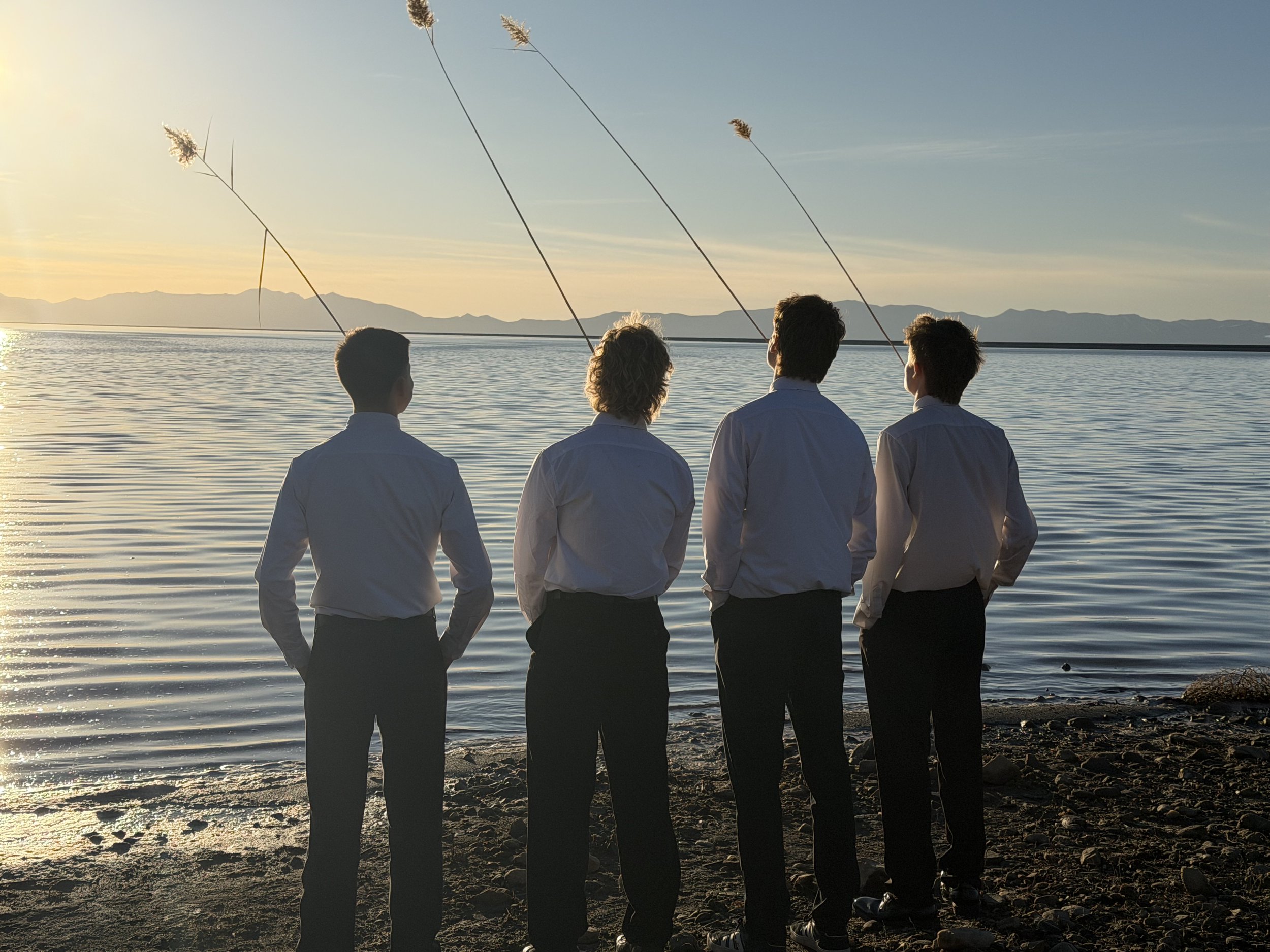 Four people dressed in white shirts and dark pants standing on a rocky shoreline, facing a body of water during sunset, with mountains in the background.