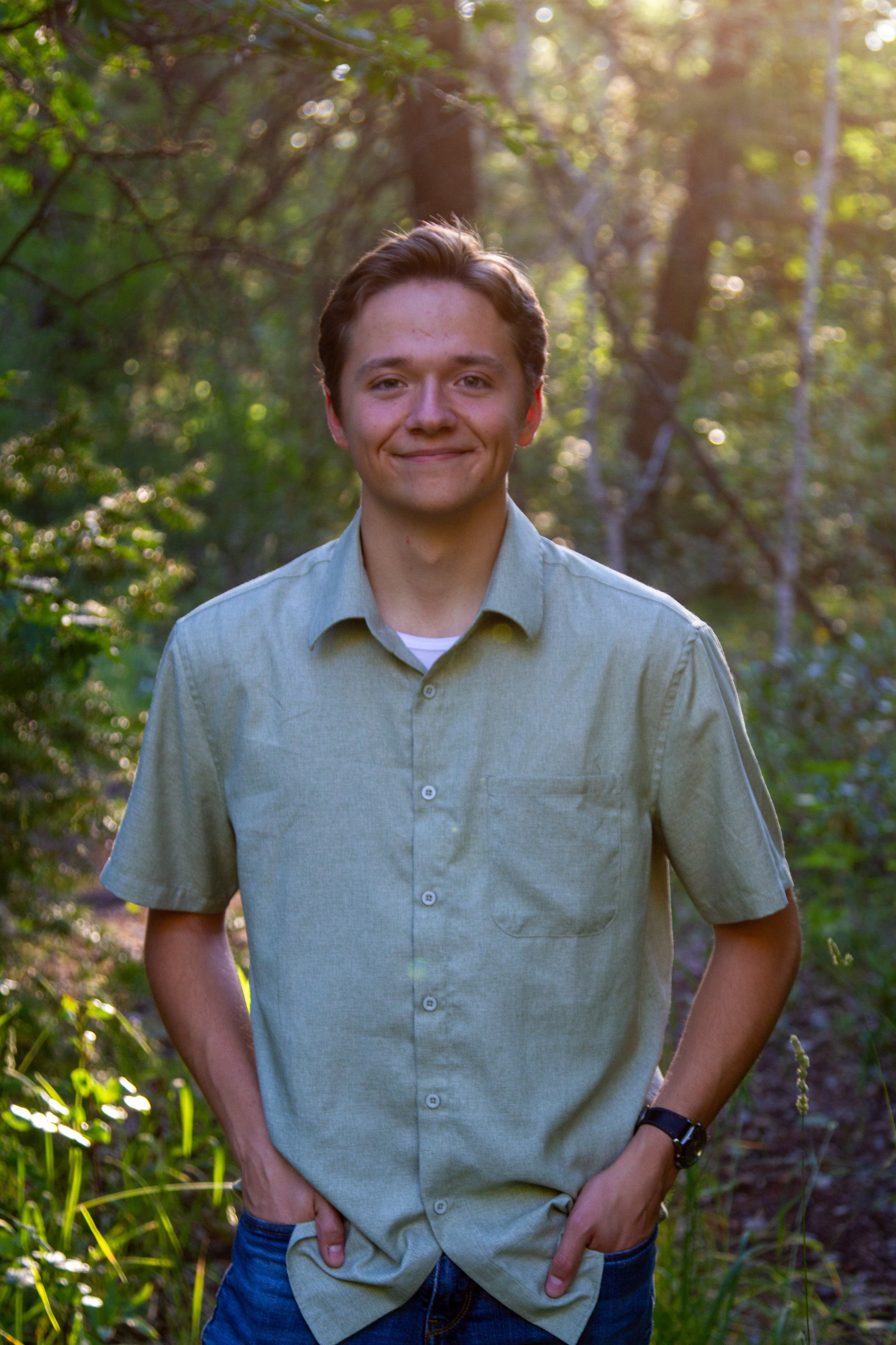 Young man standing outdoors in a forest, smiling, with sunlight filtering through trees in the background.