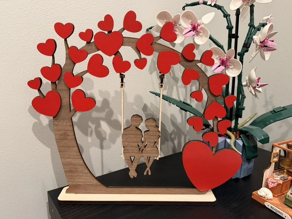 Decorative wooden and plastic art piece depicting a couple sitting on a swing under a tree with red hearts and pink flowers.