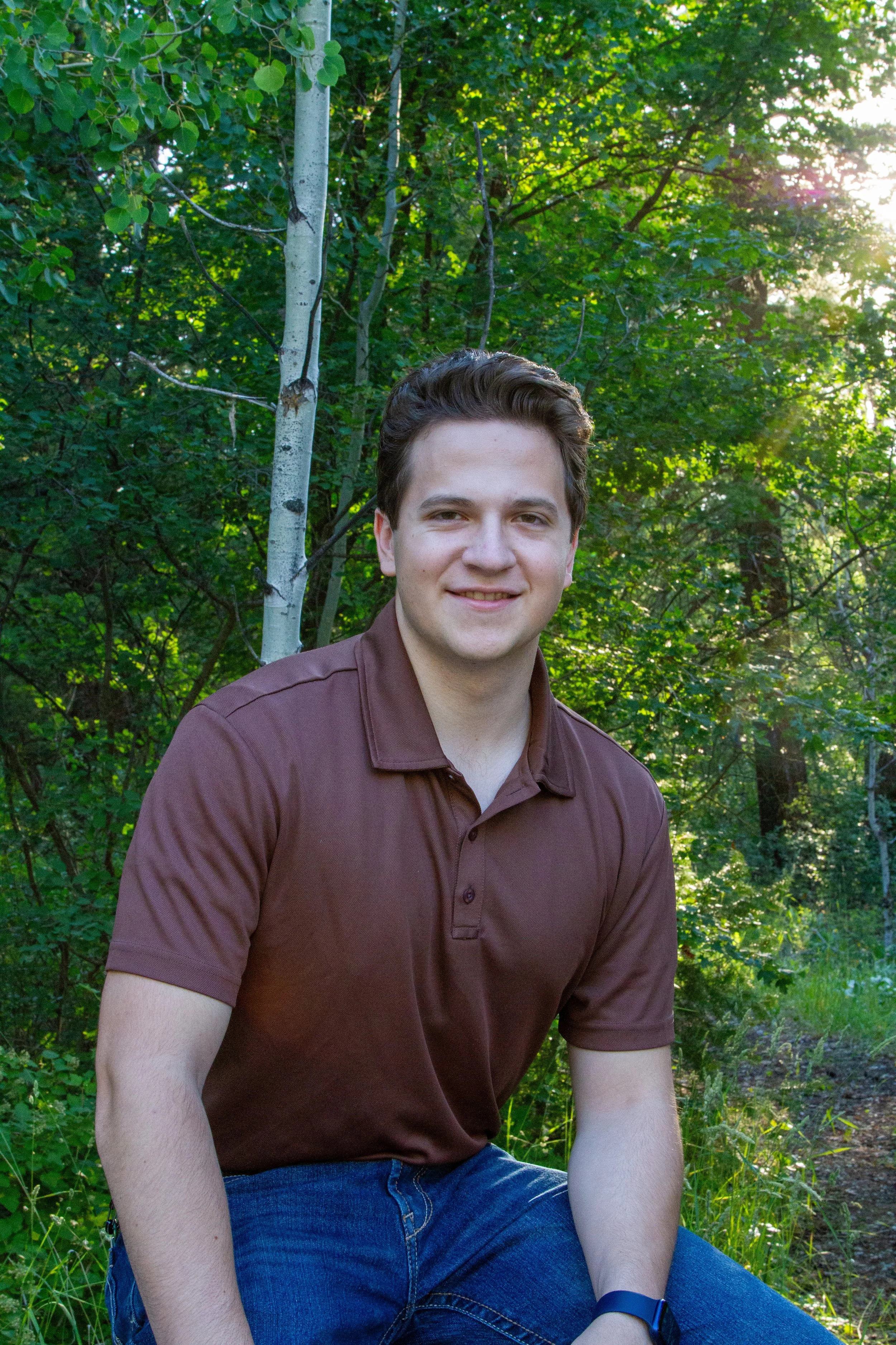 A young man with brown hair smiling while sitting outdoors in a wooded area with lush green trees and sunlight filtering through the leaves.