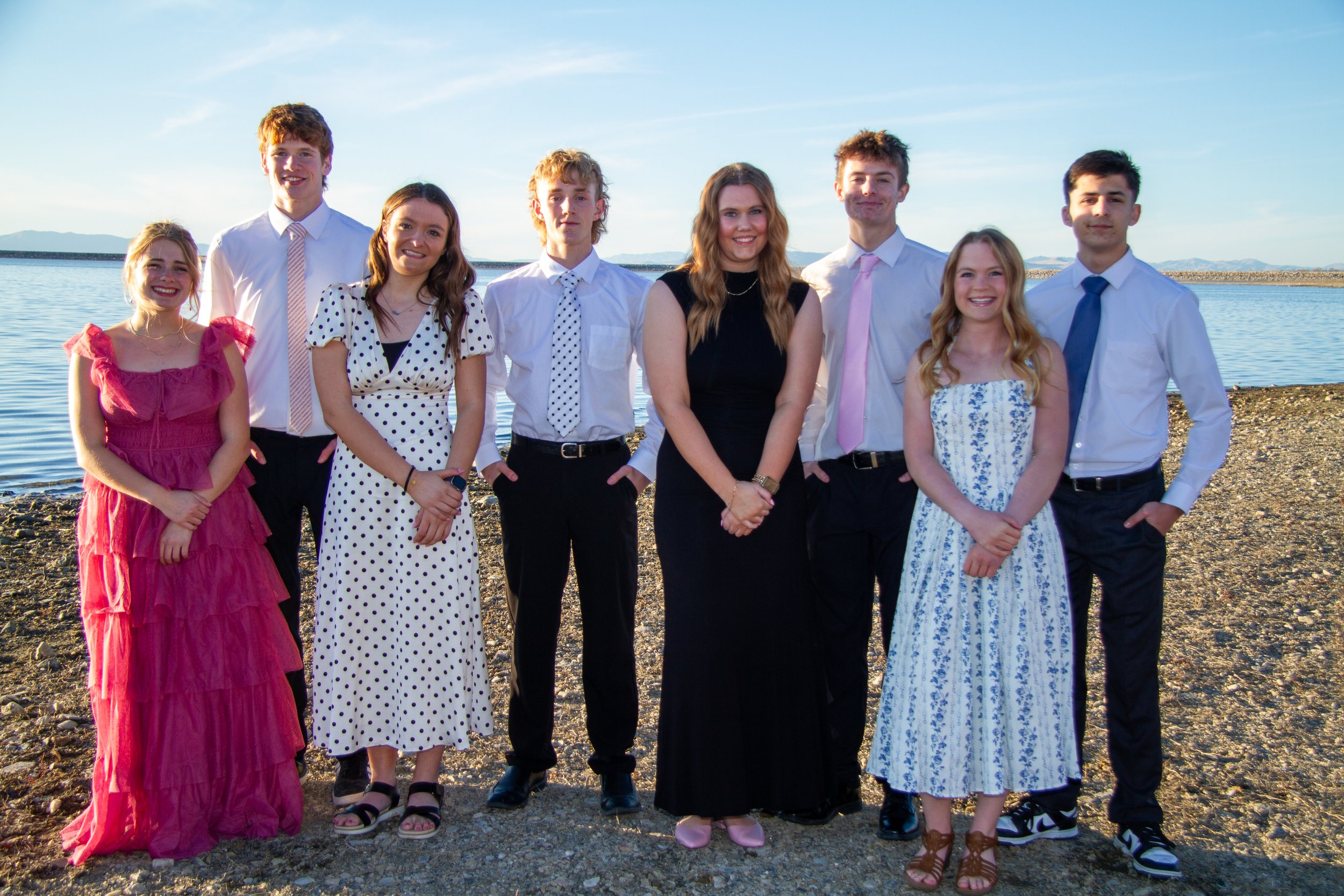 Group of nine young people in formal attire standing on a rocky beach near water with mountains in the background during daylight.
