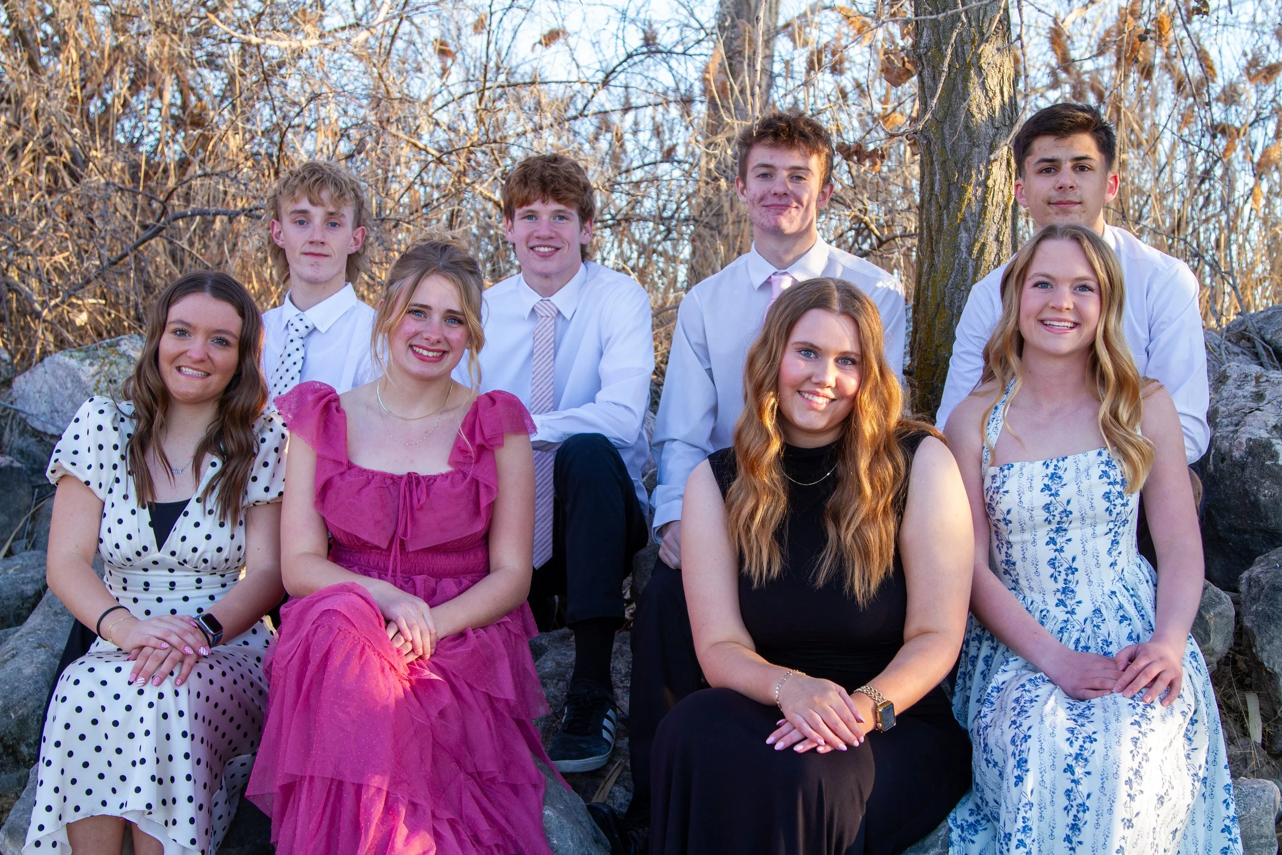 A group of eight young people sitting and standing outdoors on rocks with a leafless tree in the background during daytime.