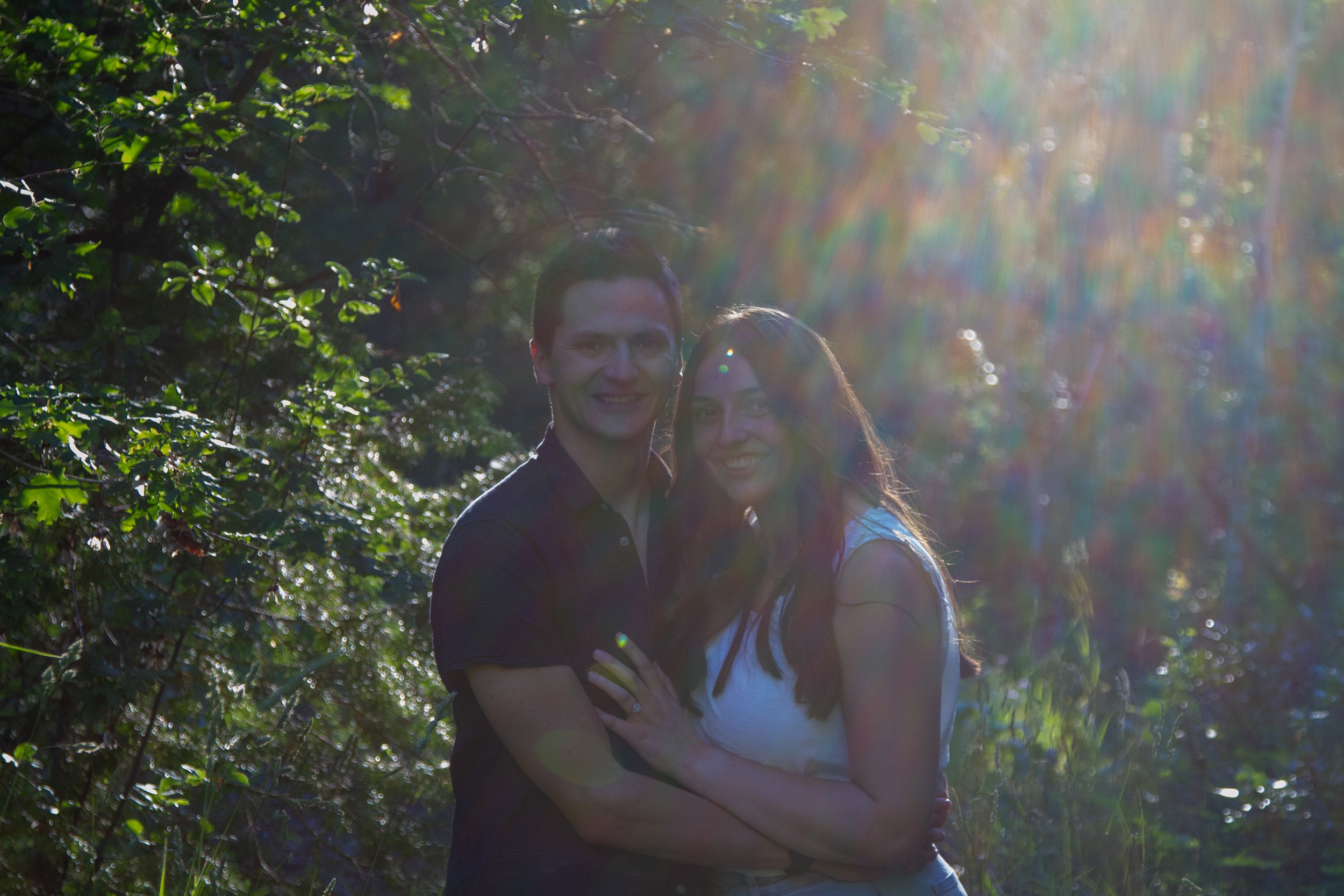 A smiling young man and woman standing close together outdoors among green trees, with sunlight creating a rainbow lens flare.