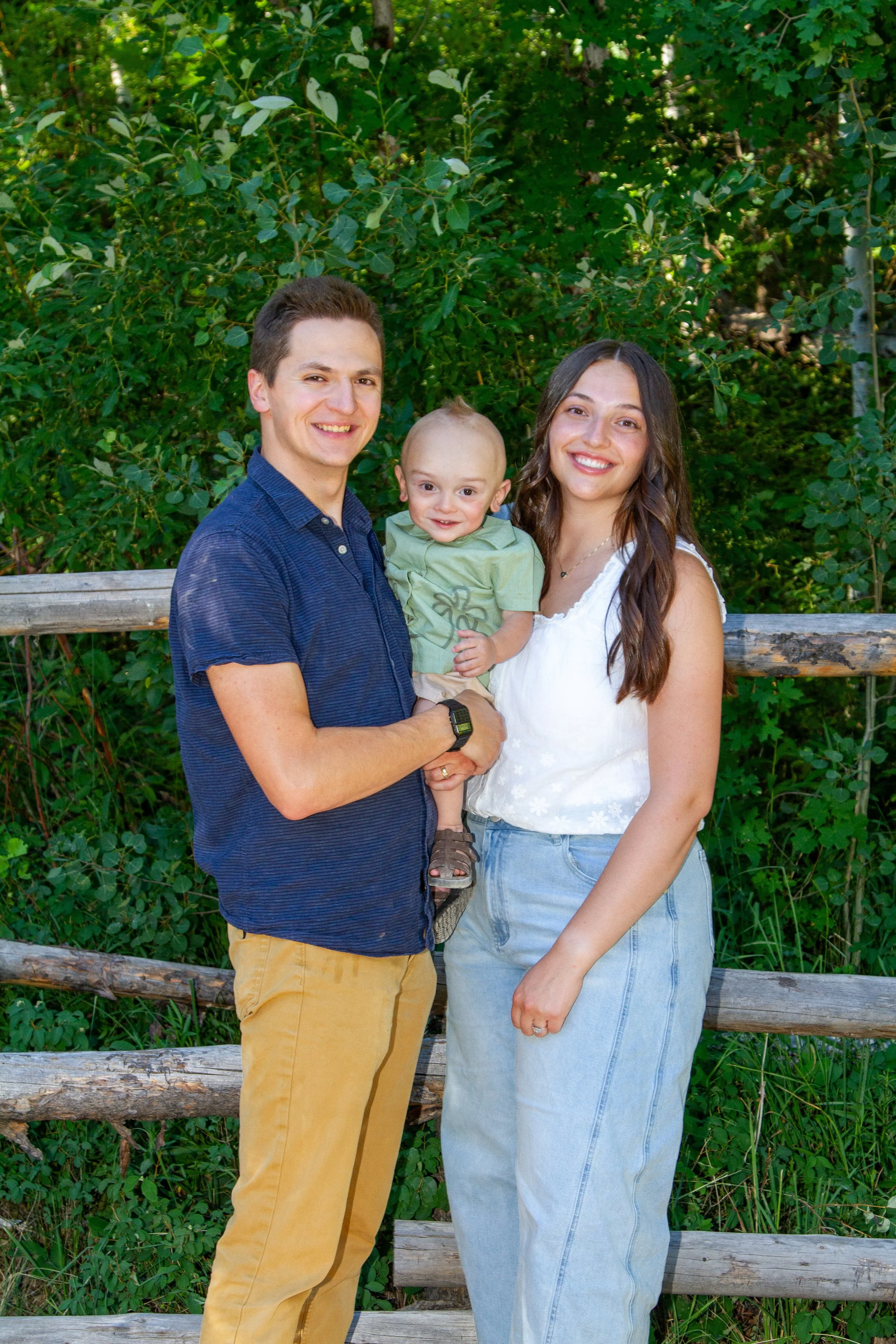 A family of three standing outdoors in front of a wooden fence and green foliage, smiling at the camera. The father is wearing a blue shirt and khaki pants, holding a young boy, and the mother is wearing a white top and light blue jeans.