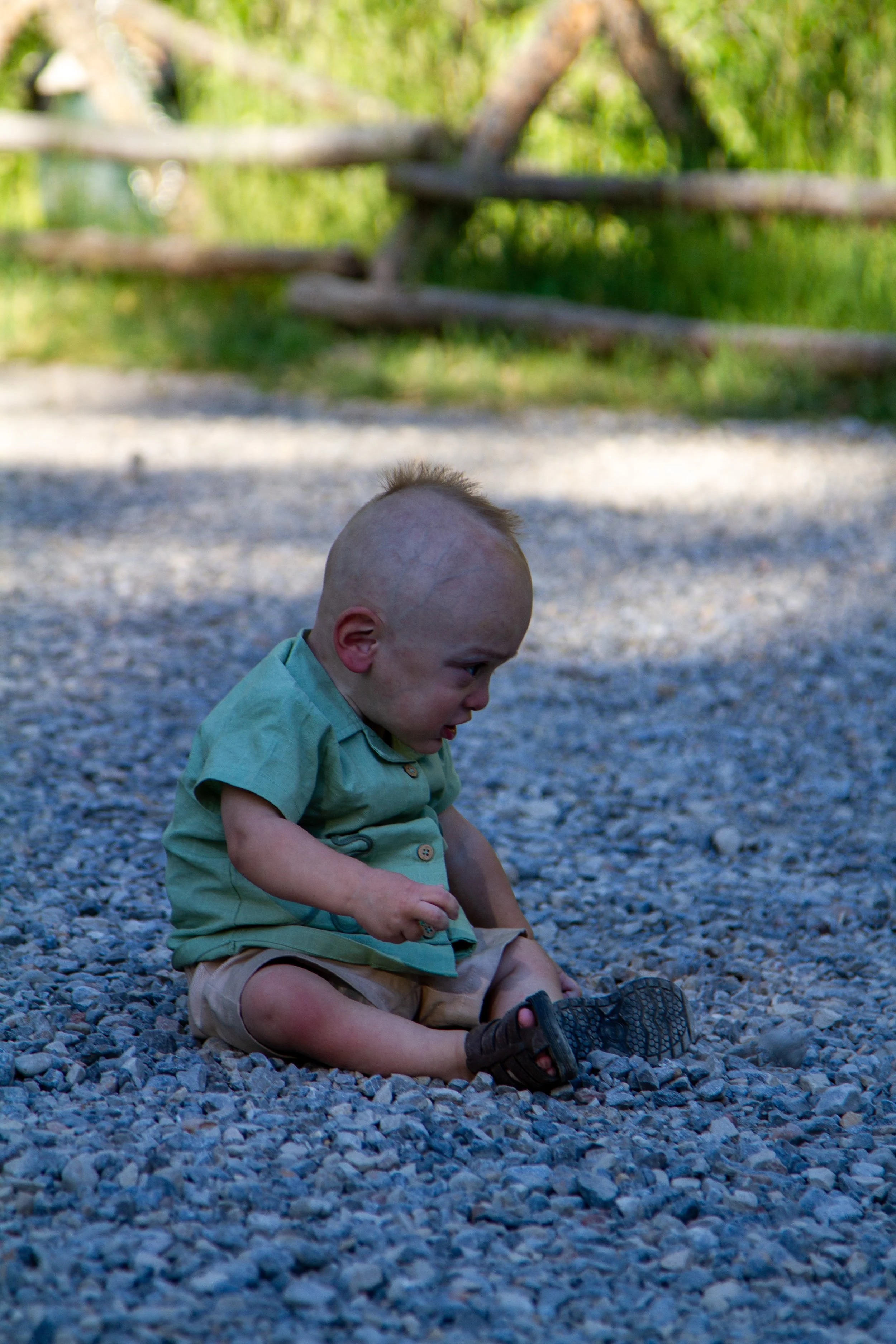 A young boy with a shaved head, wearing a green shirt and tan shorts, sitting on a gravel surface outside. There is a wooden fence and greenery in the background.