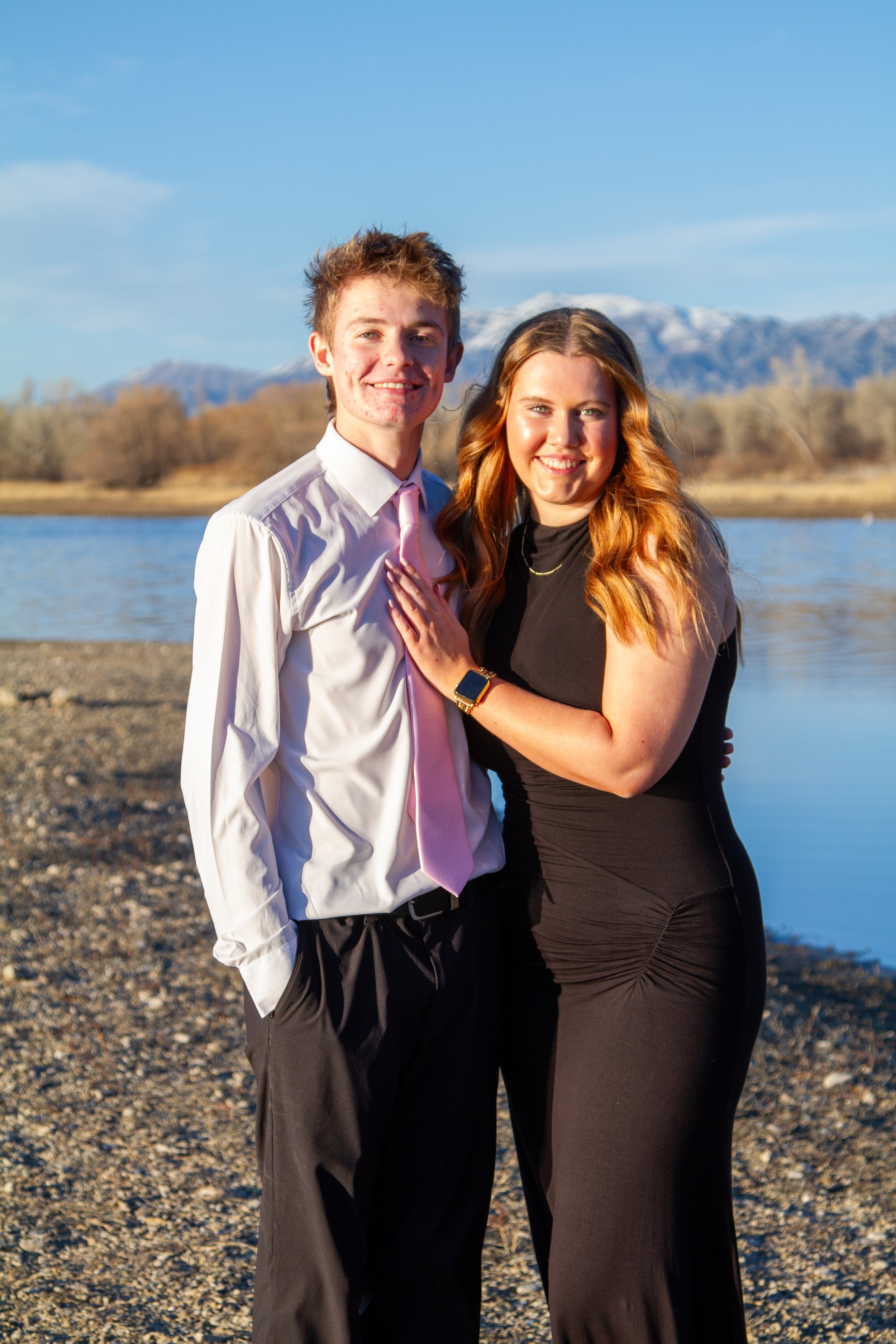A young couple standing by a lake during sunset, with mountains in the background, smiling at the camera.