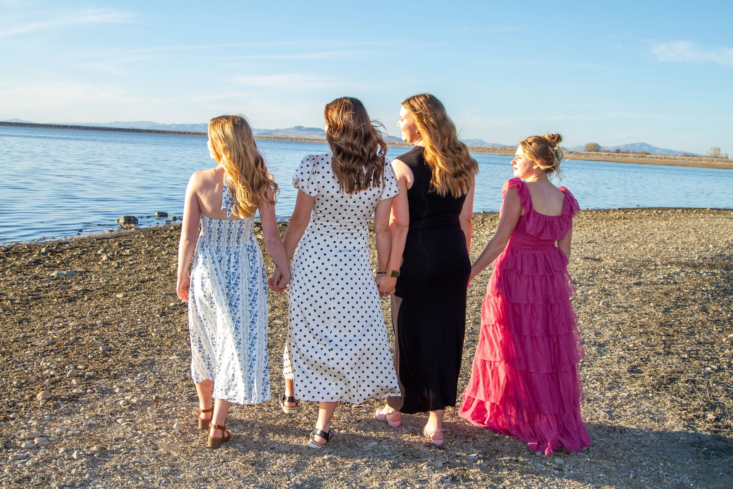 Four women wear long dresses, holding hands, walking near a body of water on a sunny day.