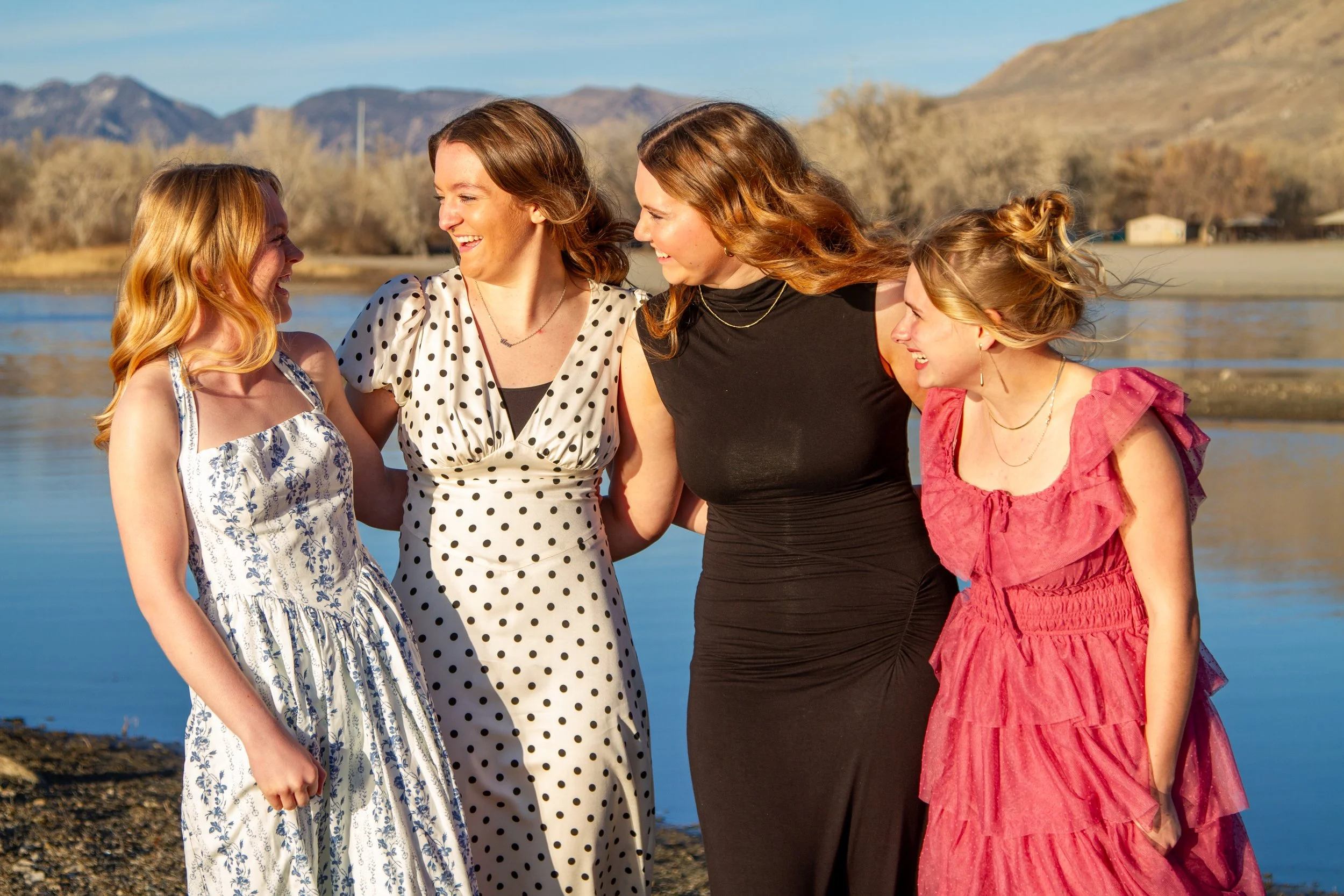 Four women standing together outdoors near a body of water, smiling and enjoying each other's company.