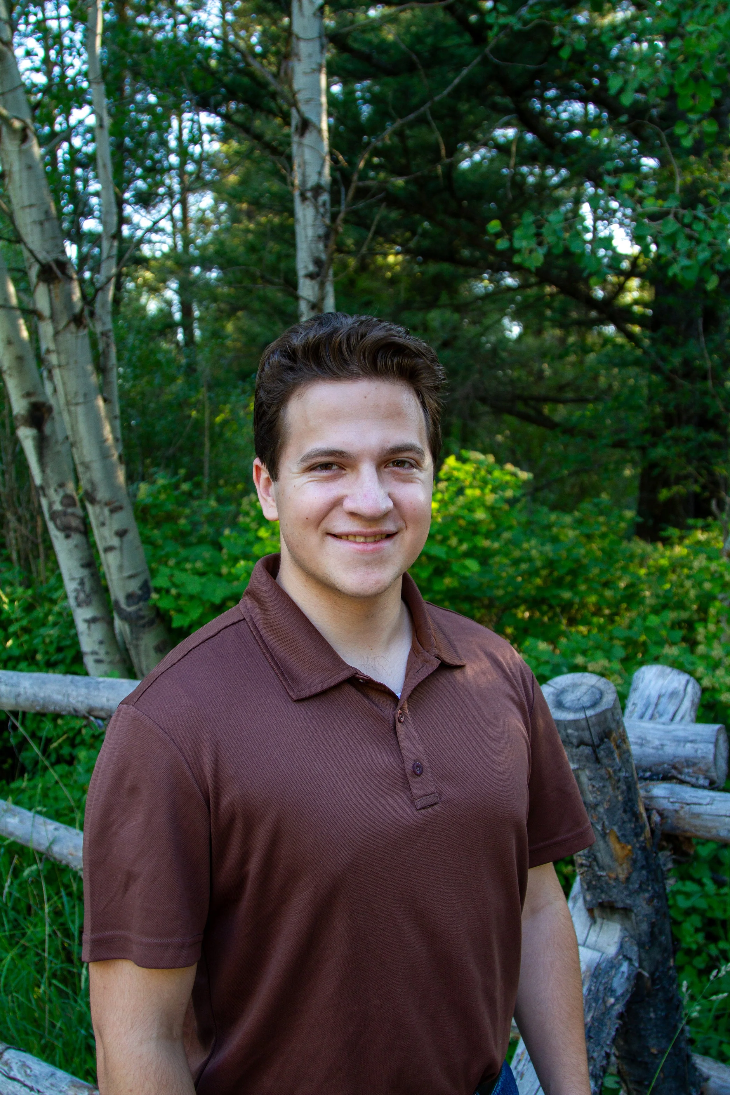 A young man with short dark hair standing outdoors in front of trees and logs, wearing a brown polo shirt, smiling at the camera.