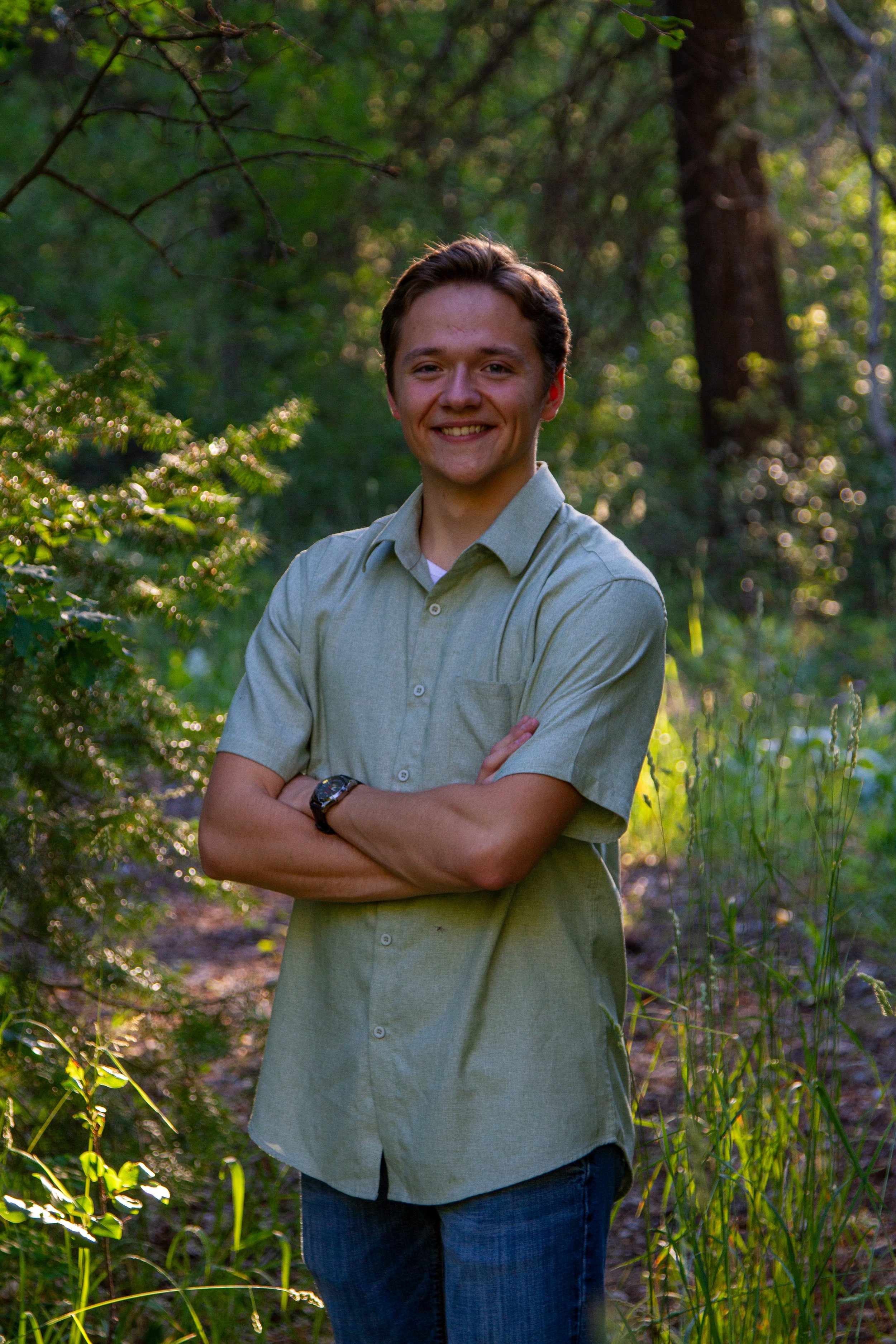 A young man smiling with arms crossed, standing in a lush green forest during daylight.