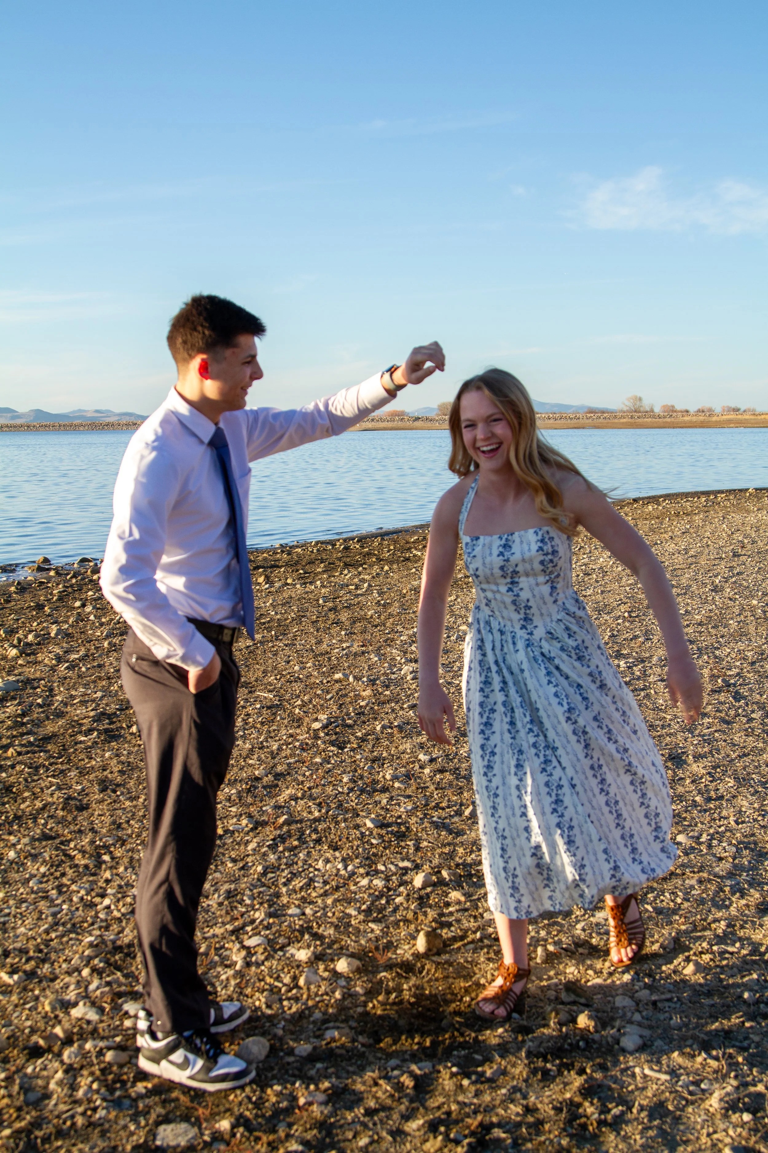 A young man and woman are dancing and laughing on a rocky shoreline near a body of water during sunset or late afternoon.