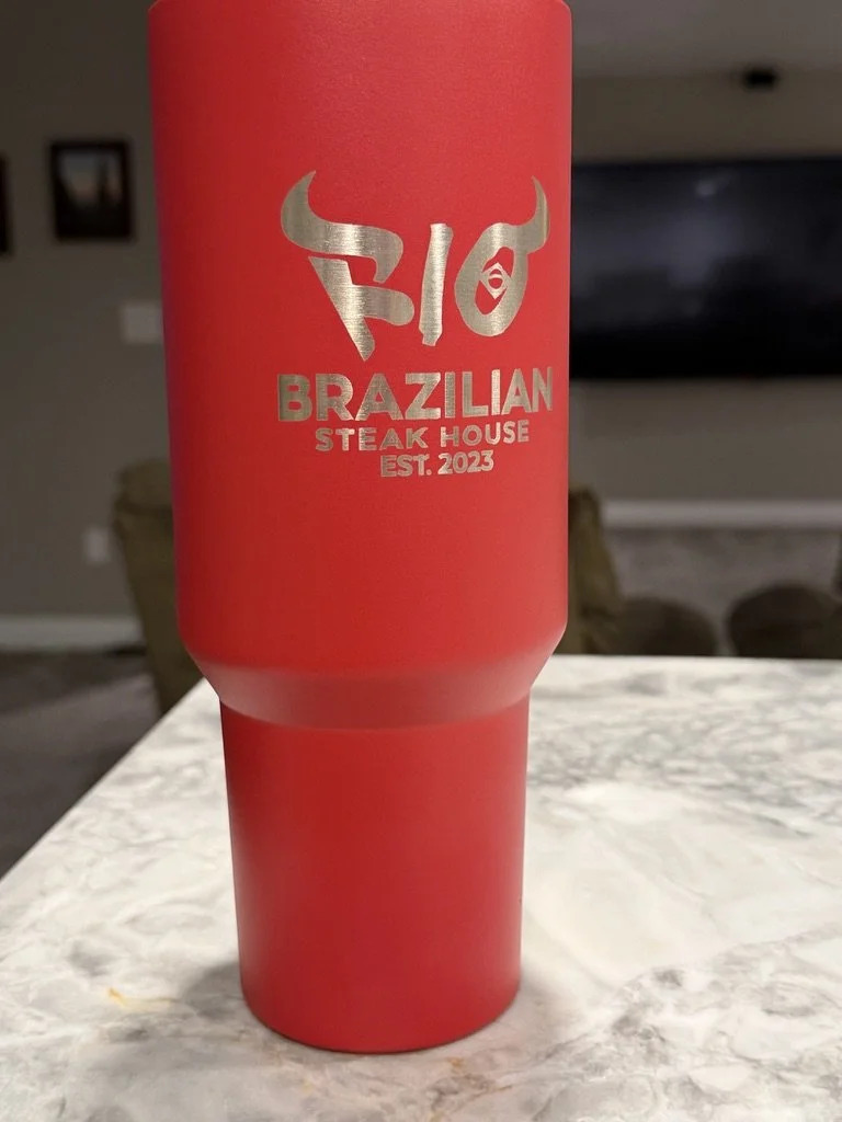 Red tumbler cup with silver logo of a bull's head and text indicating 'BIO Brazilian Steak House Est. 2023' placed on a marble countertop in a room with blurred background.