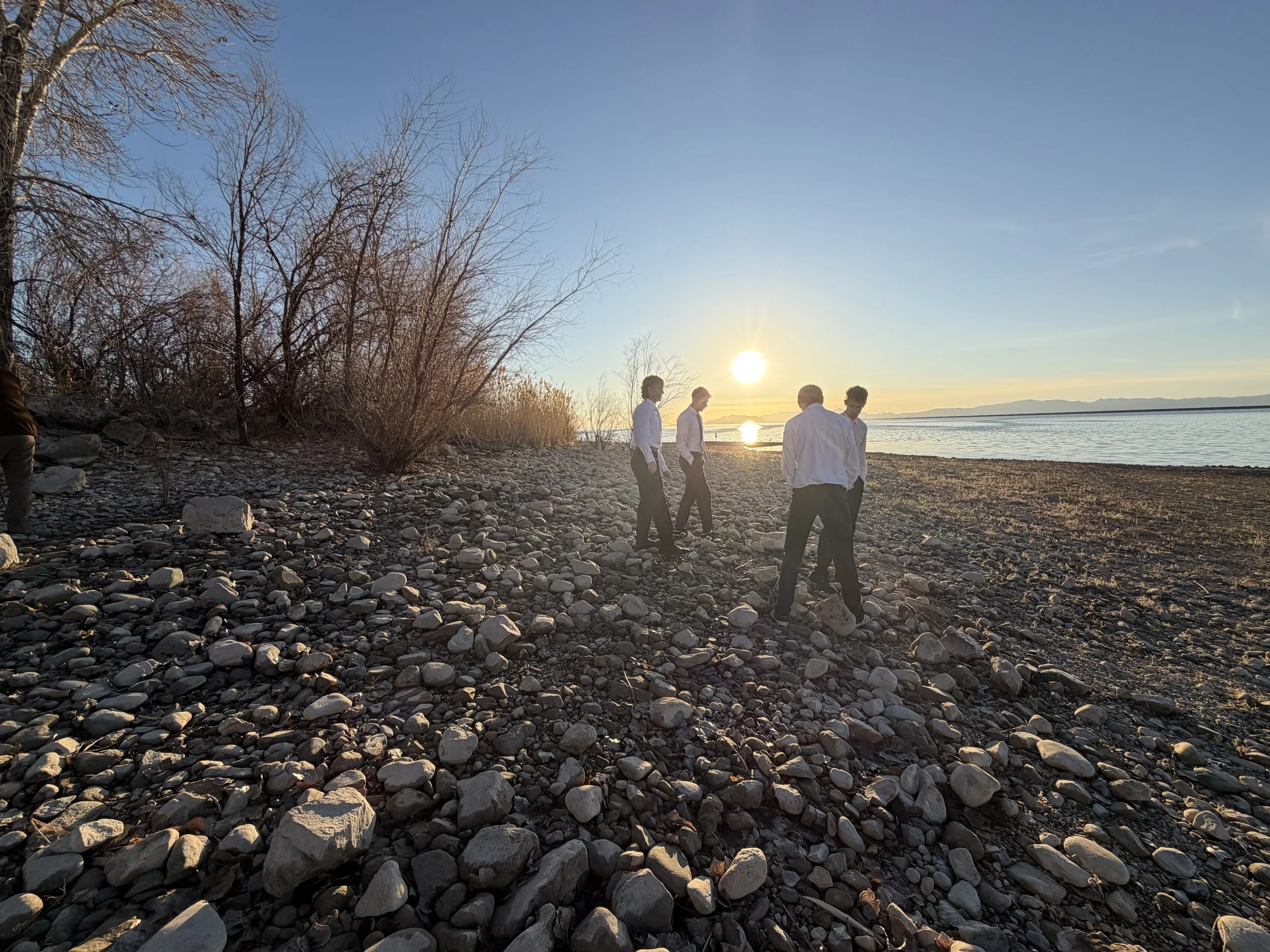 Four people in white shirts and dark pants walking on a rocky beach during sunset, with leafless trees on the left and water on the right.